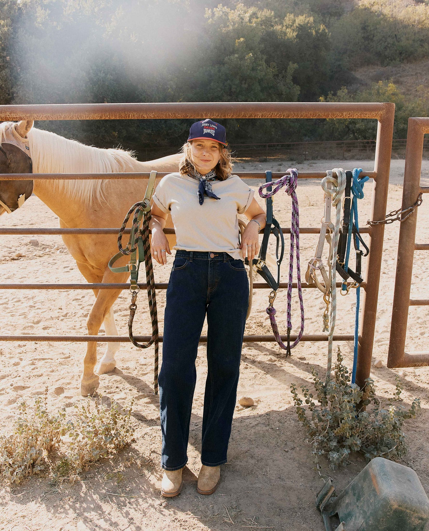 A woman stands in a dusty outdoor ranch setting next to a horse, wearing a white t-shirt, dark blue jeans, and a navy cap. She is holding ropes hanging from a metal gate, with a horse and trees in the background.