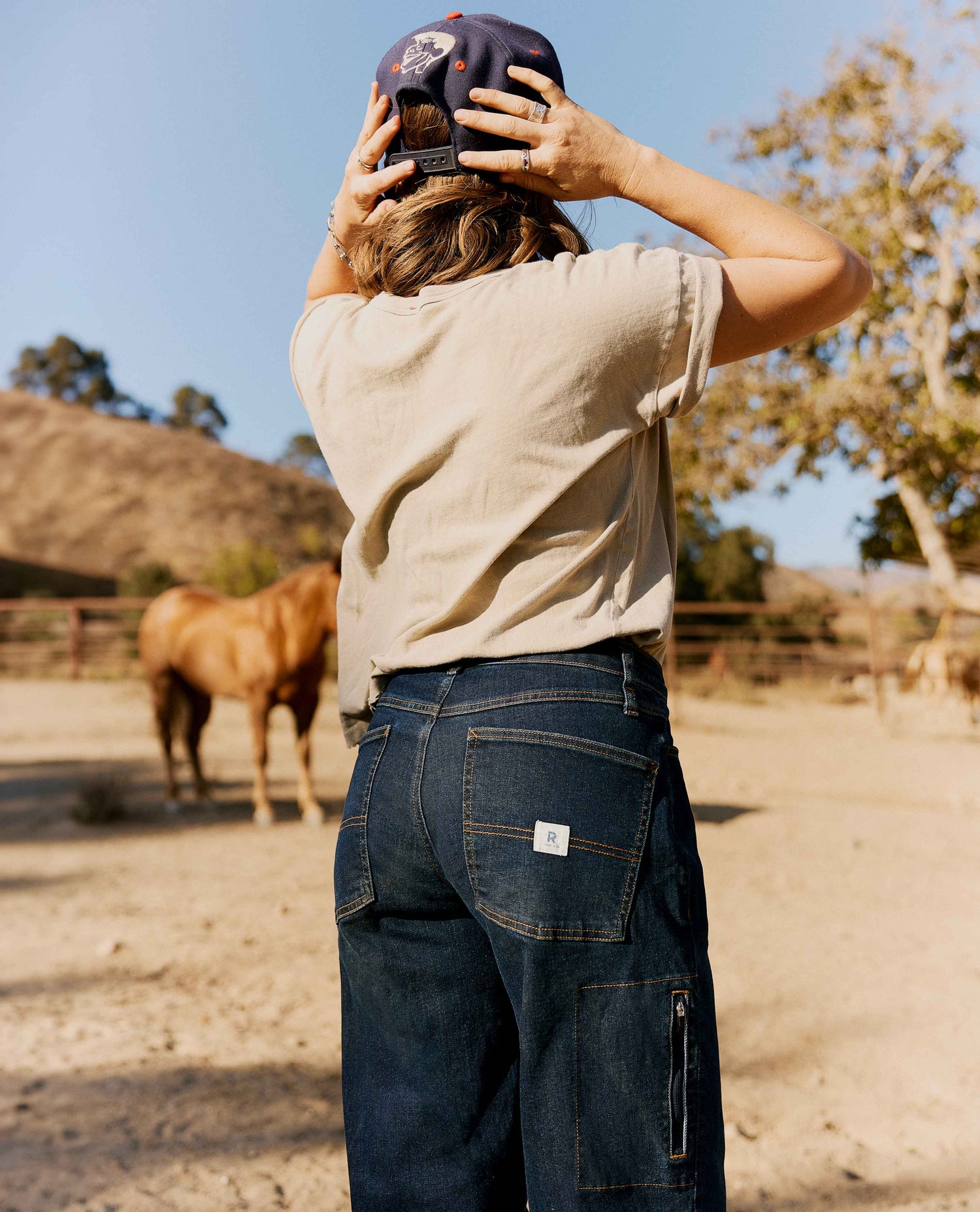 A person standing outdoors in a dusty paddock wearing a beige t-shirt, dark blue wide-leg jeans with a small white brand patch on the back pocket, and a dark cap, seen from behind.