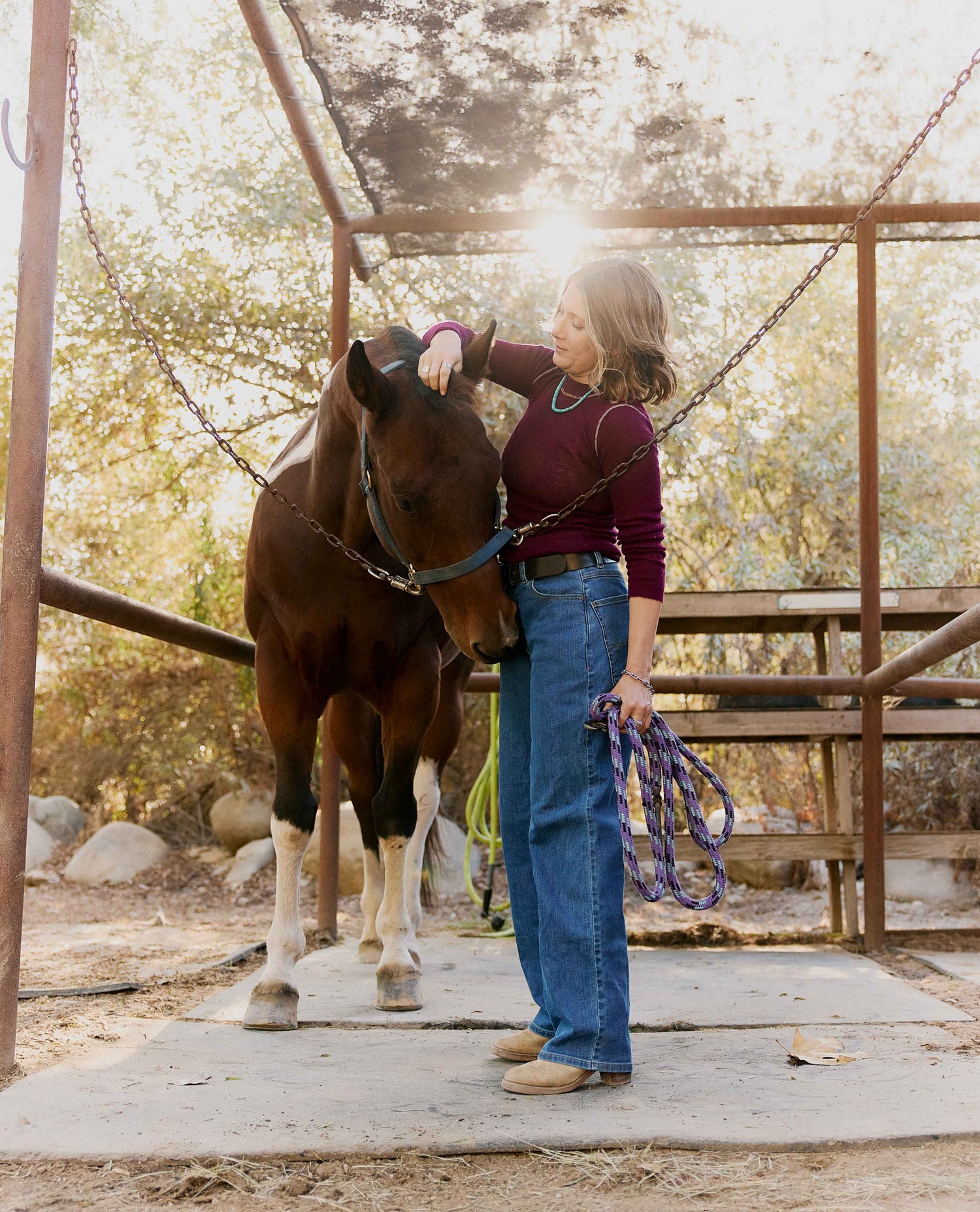 A woman in a maroon long-sleeve top and wide-leg blue jeans stands beside a brown horse in a rustic metal stable, holding a purple lead rope under warm, backlit sunlight.