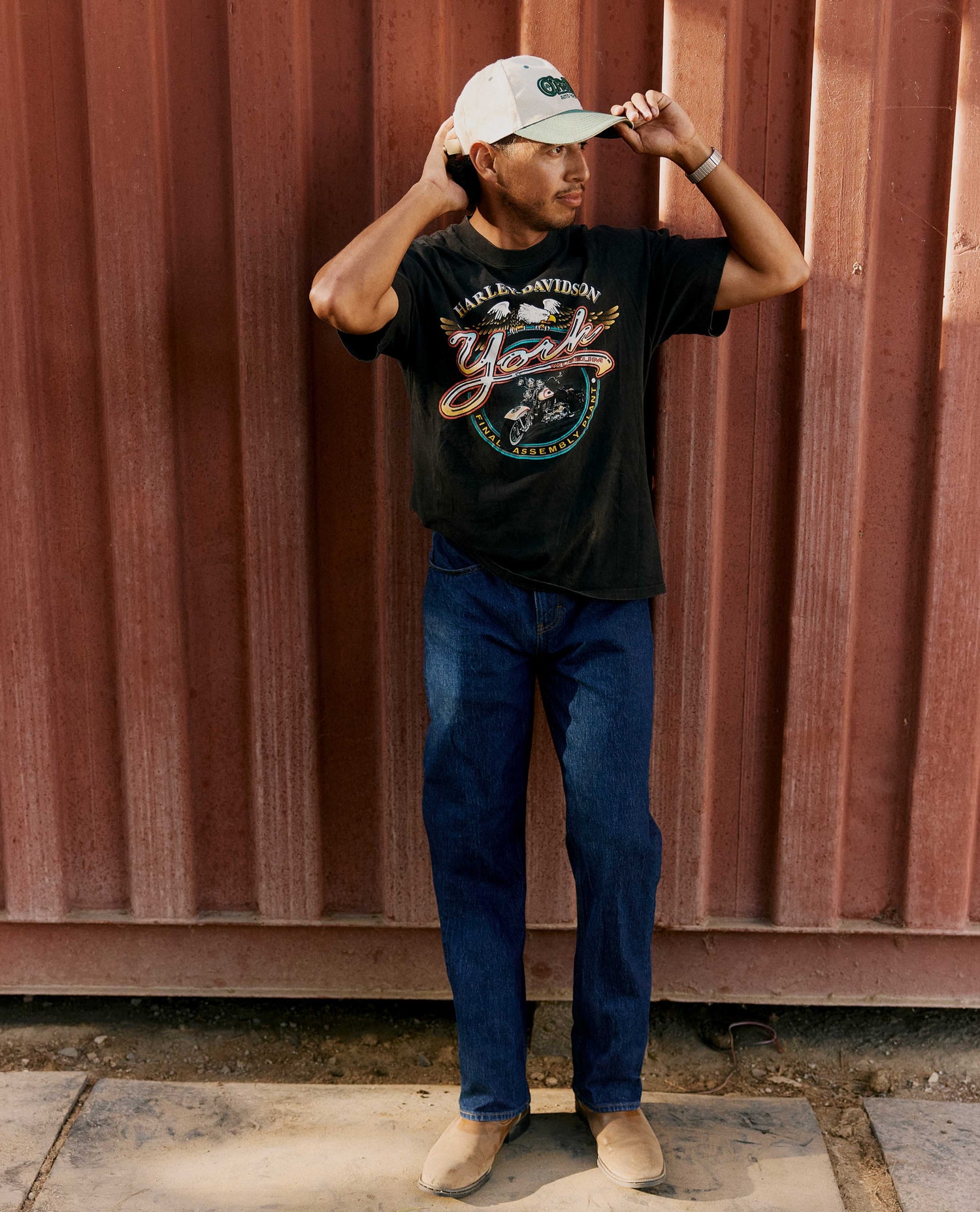 A man wearing a black graphic t-shirt, dark blue jeans, and a white baseball cap stands in front of a corrugated reddish-brown metal wall.