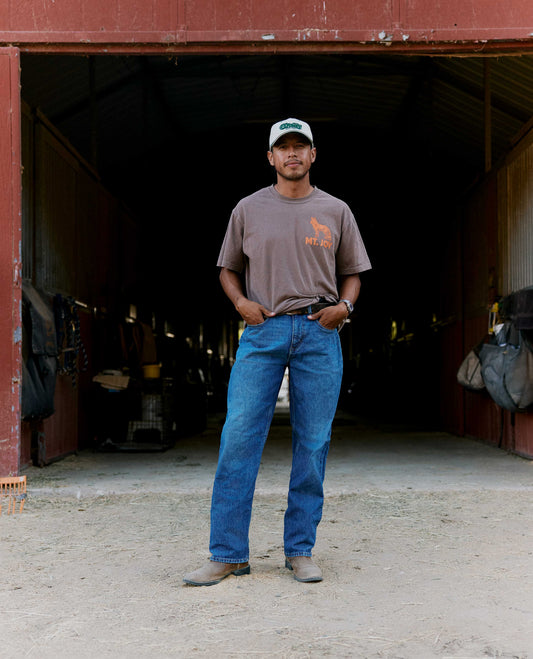 A man stands in the doorway of a rustic barn wearing a brown t-shirt with an orange graphic, blue jeans, and a white cap.