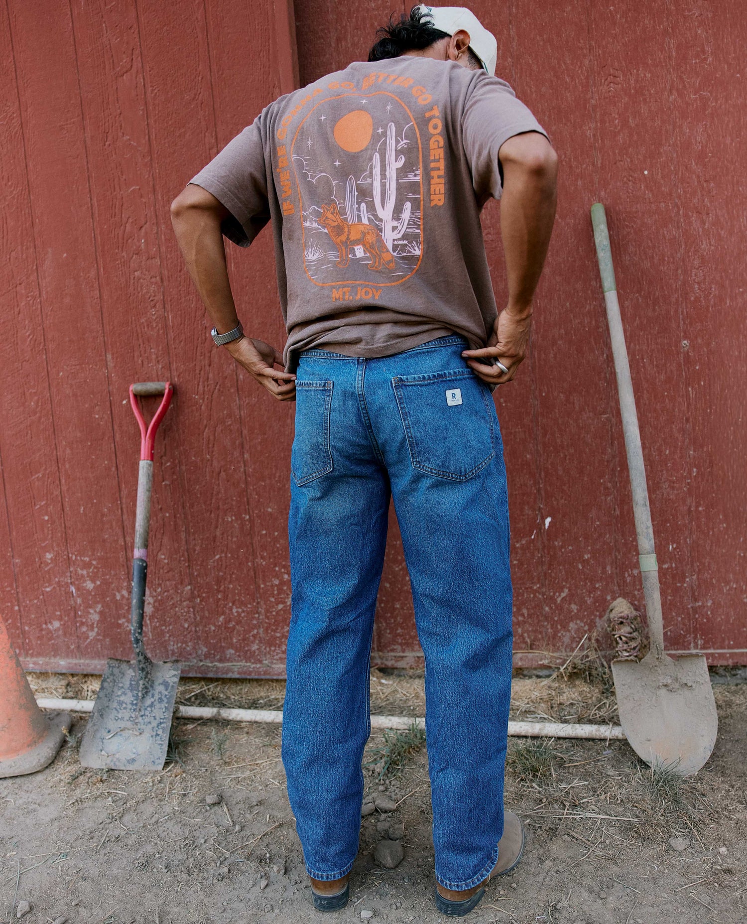A man stands with his back to the camera wearing a brown graphic t-shirt and blue jeans in front of a red wooden barn wall with shovels.