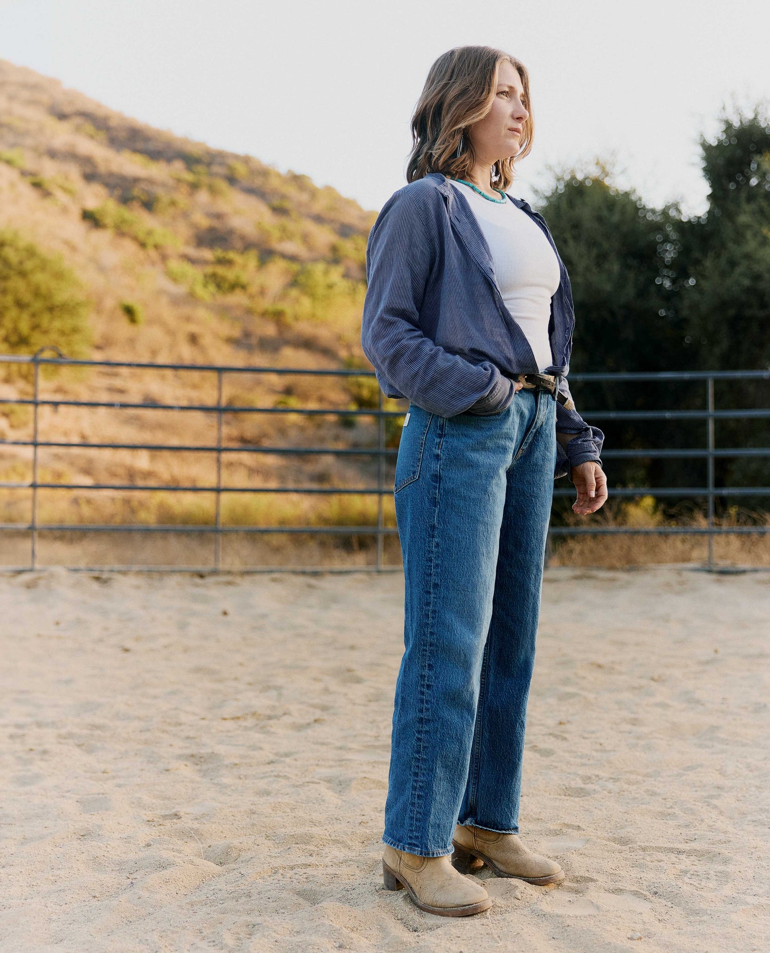 A woman stands outdoors in a sandy area with hills and trees in the background, wearing a navy blue cardigan over a white top, blue jeans, and tan boots.