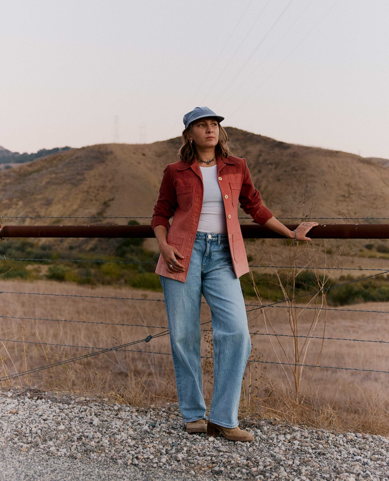 A woman stands outdoors in a dry, hilly landscape wearing a rust-red denim jacket over a white top, light-wash wide-leg jeans, and a blue cap.