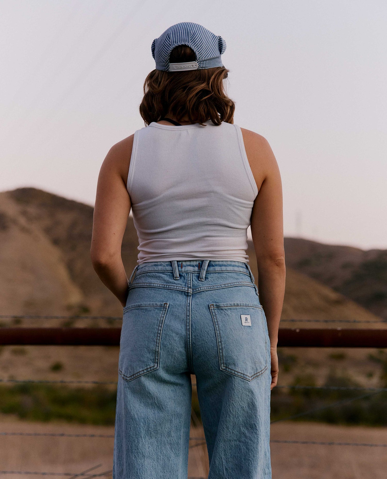 A person viewed from behind wearing a white ribbed tank top, light blue wide-leg jeans with a small white brand patch on the back pocket, and a blue-and-white striped baseball cap, standing outdoors in front of hills.