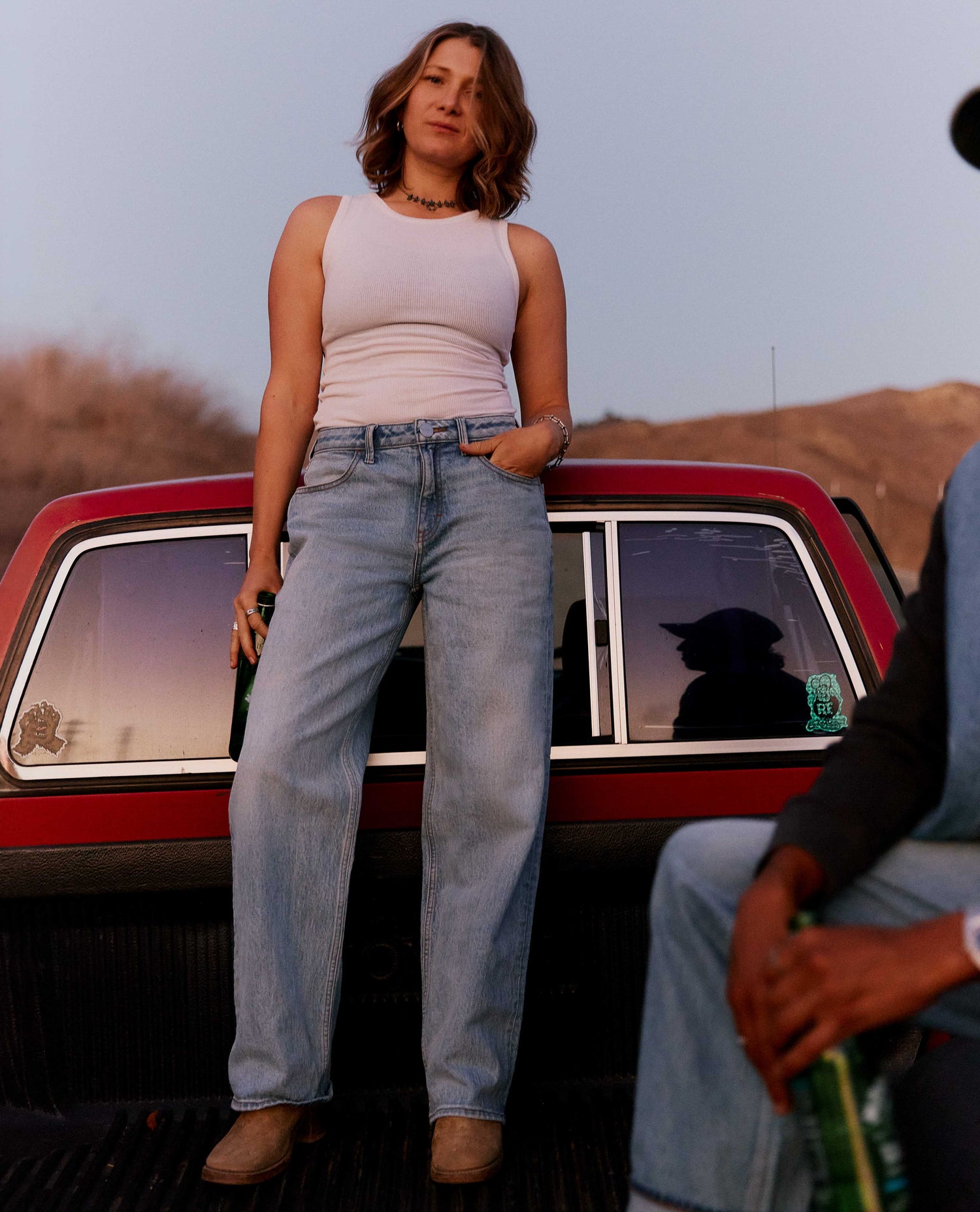 A woman stands in the bed of a red pickup truck wearing a light pink ribbed tank top and light-wash blue straight-leg jeans. A man is partially visible sitting on the right side of the truck bed.