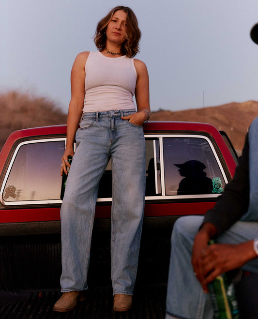 A woman stands in the bed of a red pickup truck wearing a light pink ribbed tank top and light-wash blue straight-leg jeans. A man is partially visible sitting on the right side of the truck bed.