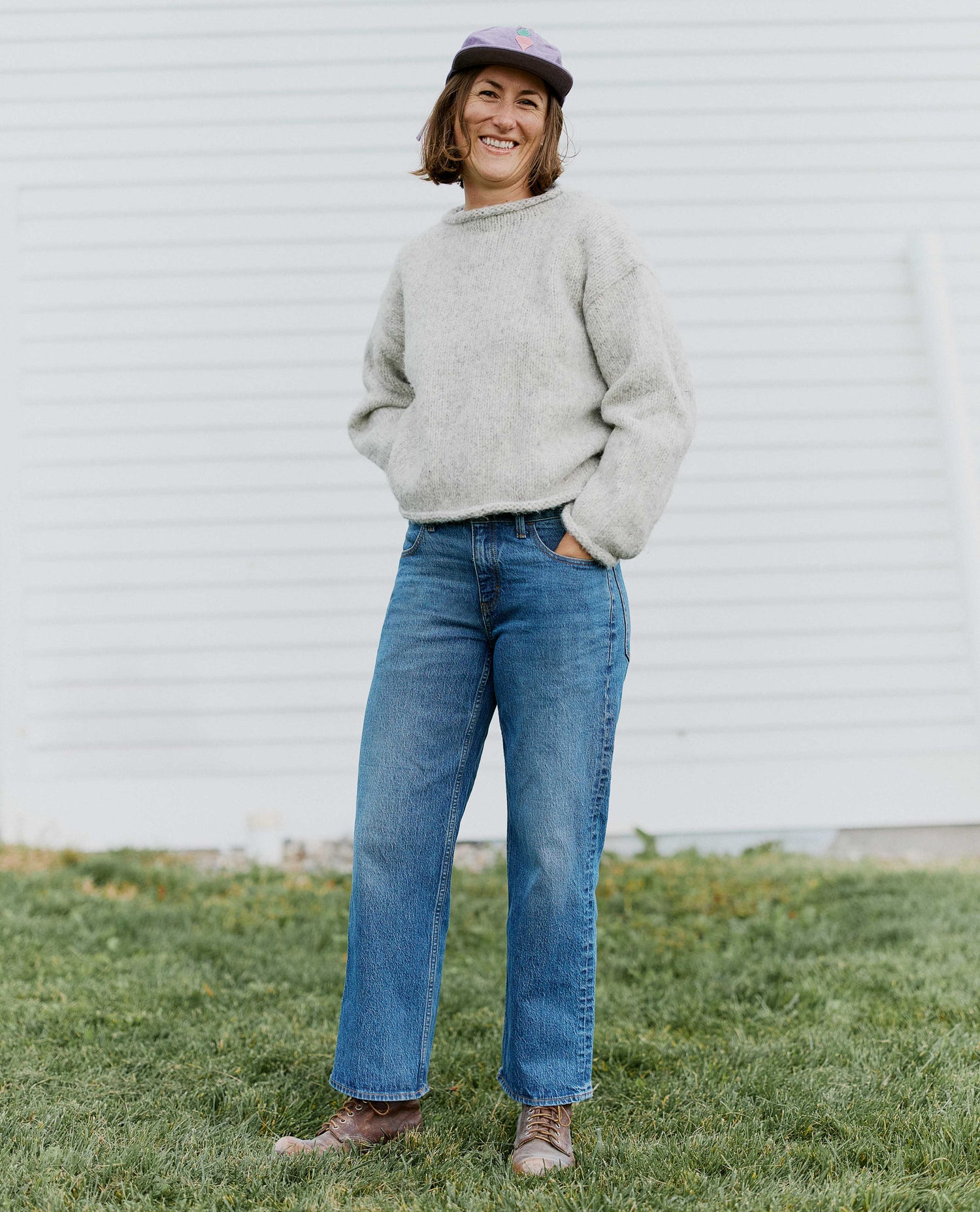 Woman stands in front of a white barn wearing medium wash blue jeans, a gray sweater, purple hat, and brown boots. 
