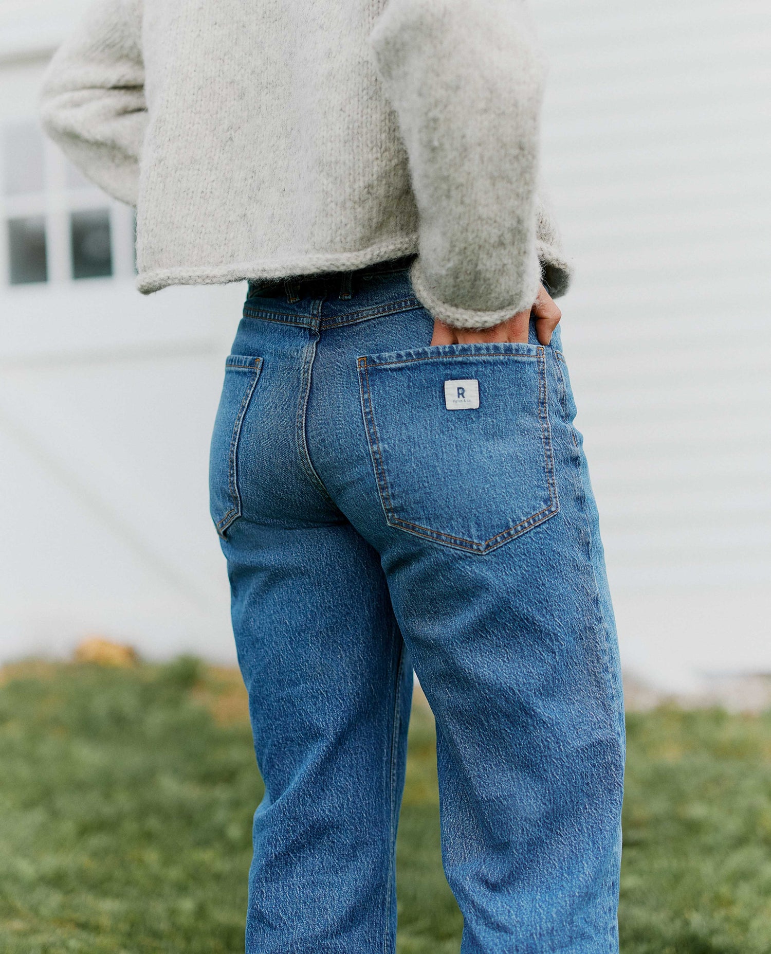 Close up of medium wash blue jeans from behind, woman wearing gray sweater with hand in back pocket of jeans. 