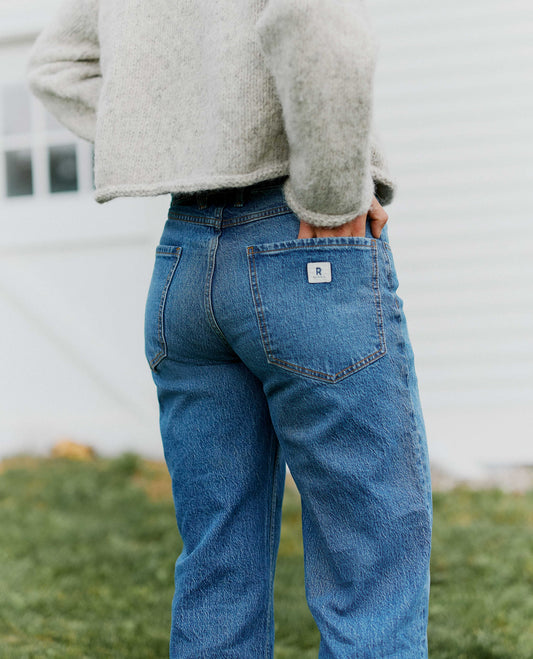 Close up of medium wash blue jeans from behind, woman wearing gray sweater with hand in back pocket of jeans. 