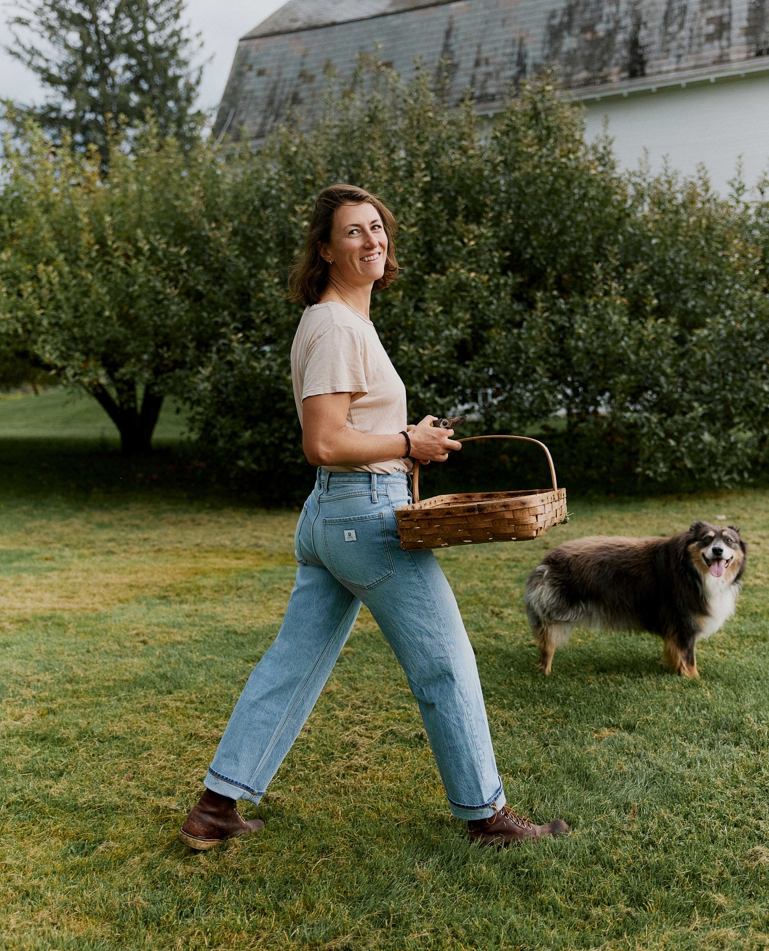 Woman wearing light wash blue jeans, holding a wicker basket and walking in front of a bush with a dog in the background.