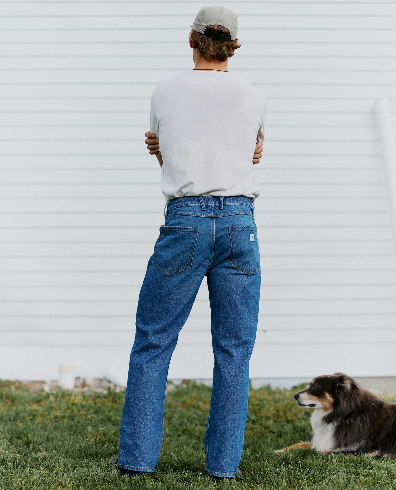 Man facing away from the carmera with jeans and a white shirt with a dog laying in the grass