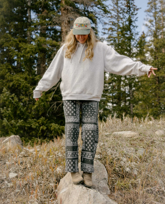 Woman wearing gray hoodie and black and white patterned pants. She is standing on a rock with boots and a baseball hat. 