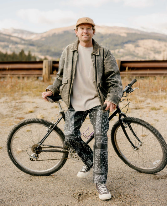 Man leaning against a bike with mountains in the background. He is wearing a gray jacket, brown hat, tan shirt, and black and white patterned pants. 