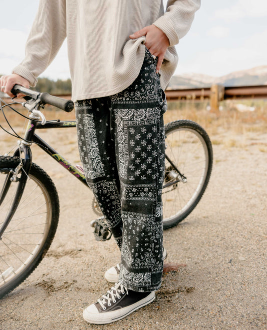 Close up of man holding a bike, wearing a tan shirt and black and white patterned pants. He has black and white shoes on. 