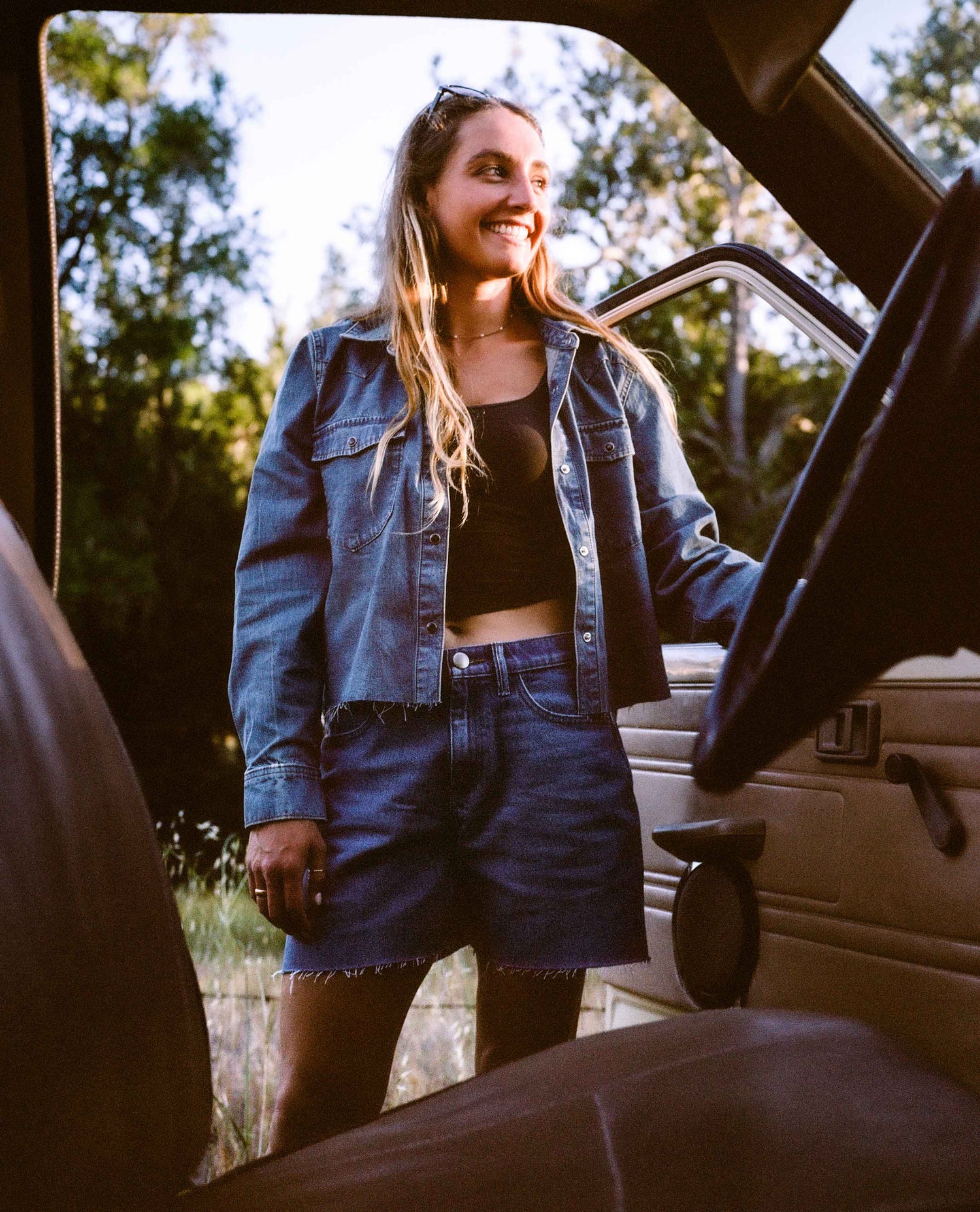 A woman smiling and looking out from inside a vehicle, wearing a cropped blue denim shirt and denim shorts over a black crop top.