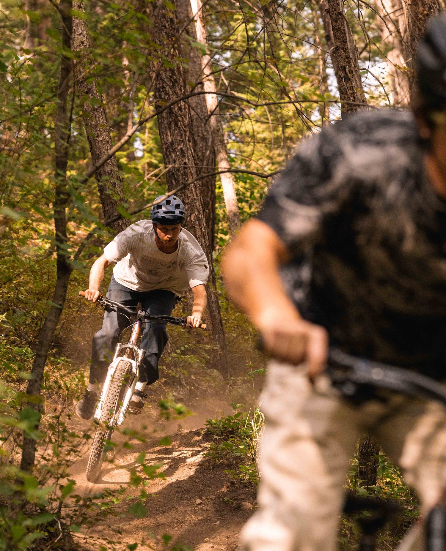 Two men ride their mountain bikes in the woods. The first man is out of focus. He is wearing a black and grey shirt with white pants. The second man is in focus. He is in a blue helmet, grey t shirt, and blue pants. 