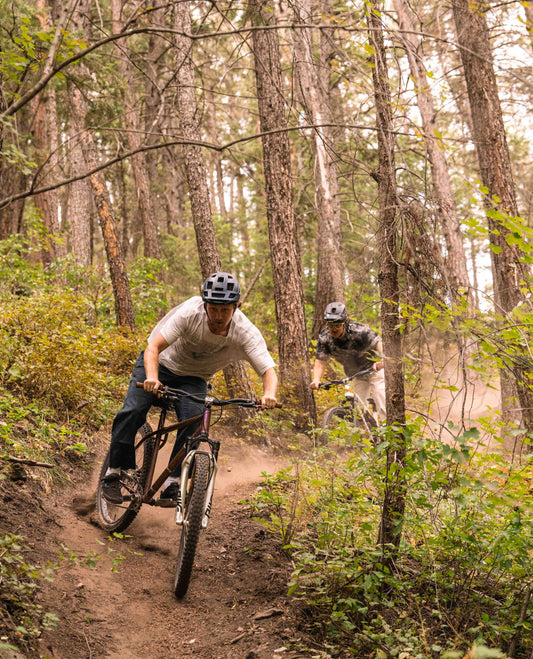 A man riding his mountain bike through a corner in the woods. He is wearing a helmet, a blue jacket and pants, and a grey t shirt. The jacket flaps behind him as his rear wheel scatters dust. A second man follows him. Second man is wearing a helmet, glasses, a black and grey shirt, and white pants.