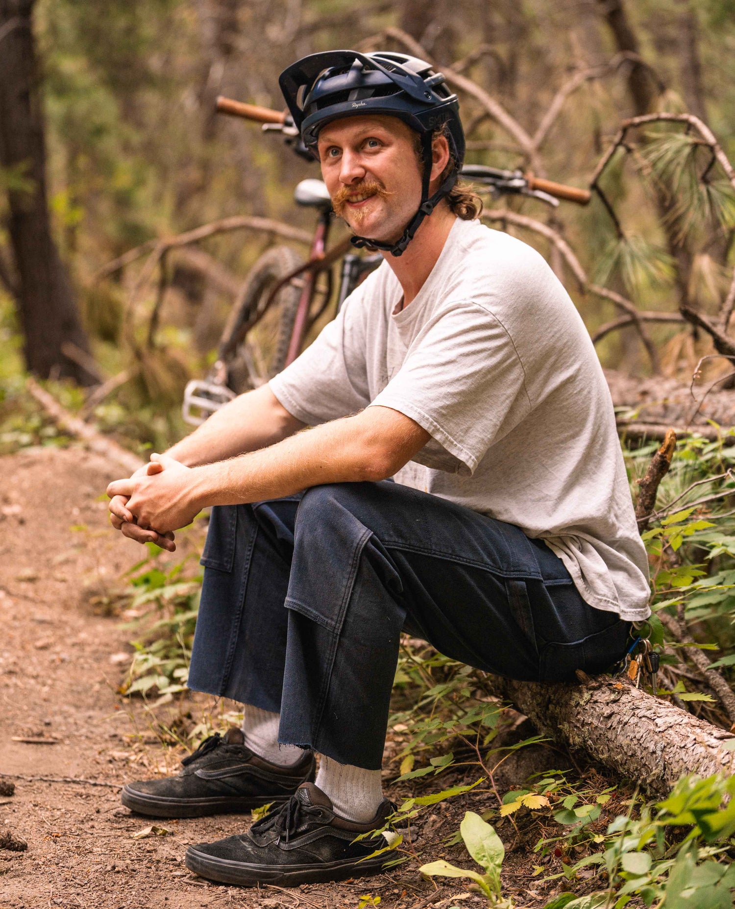 A man sitting on a log in the woods. He is wearing a blue helmet, white t shirt, and blue argo pants with black shoes.