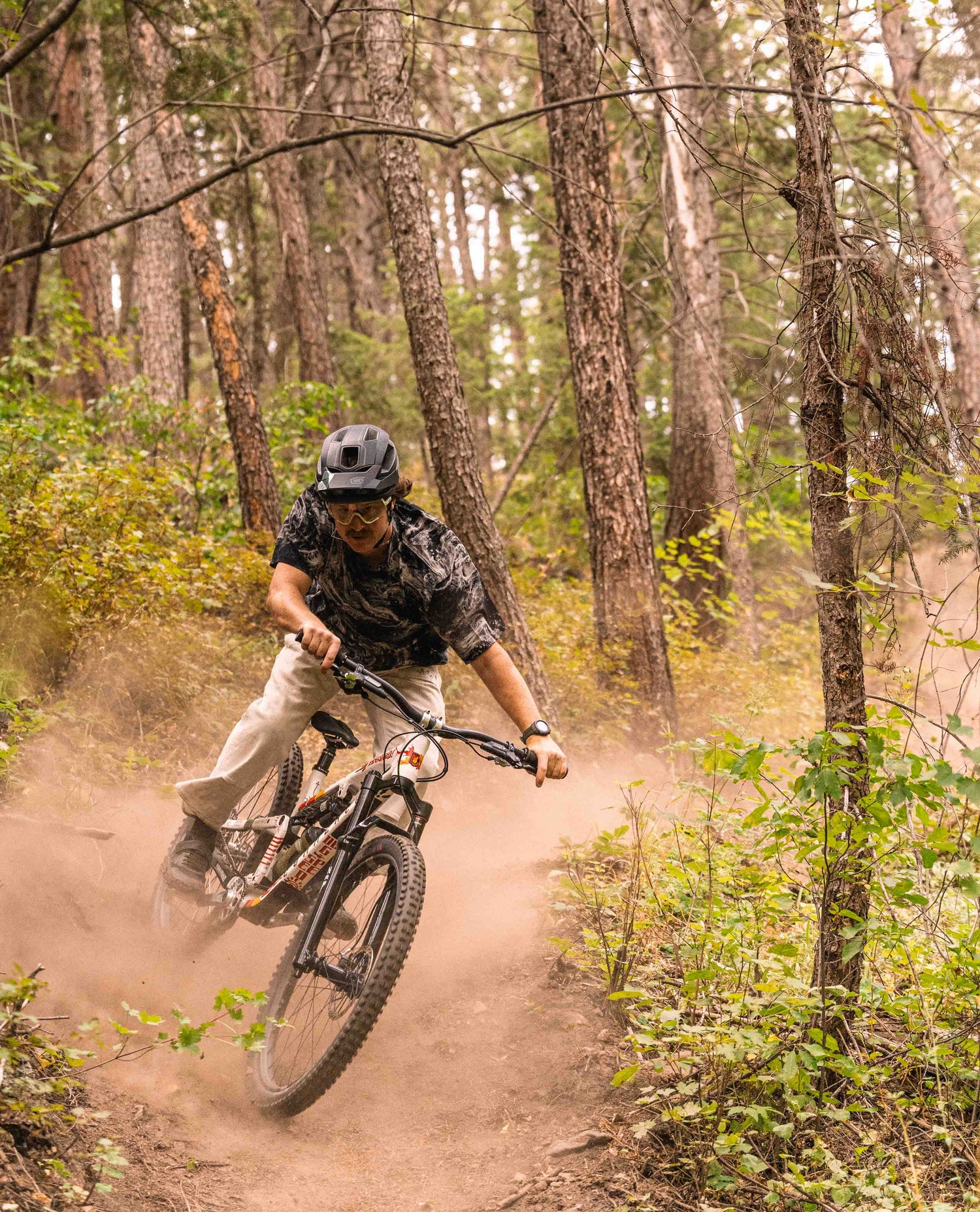 A man riding his bike through a corner in the woods. He is wearing a helmet, glasses, a marbled grey and white jersey, white pants, and black shoes. His bike is white with graffiti.