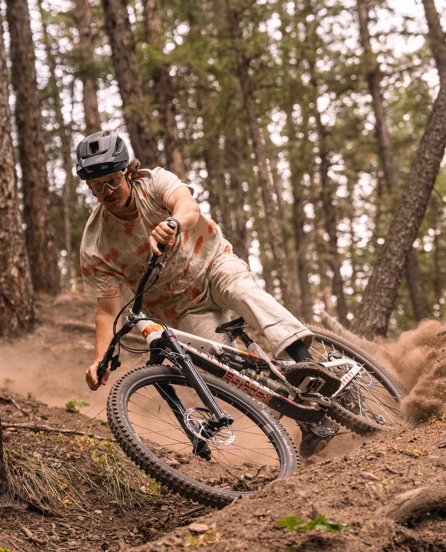 A man riding through a corner on his mountain bike. He is spraying dust behind him. The rider is wearing a helmet, glasses, white and orange speckled t-shirt, as well as white pants. His bike is white with graffiti.