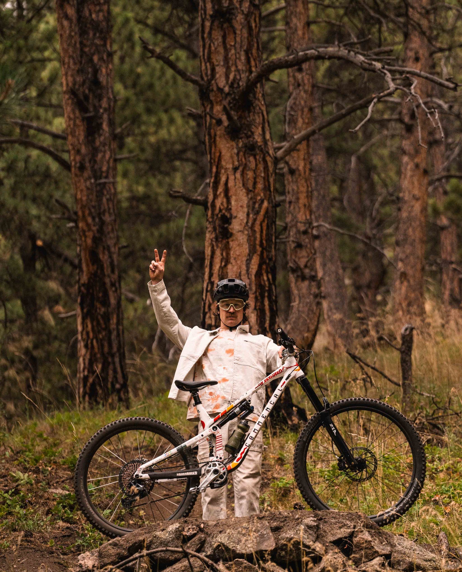 A man standing with his mountain bike in front of a large tree within a forest. He is gesturing a "peace" sign. He is wearing a helmet, grey and yellow glasses, white jacket and pants and a white and orange speckled shirt. His bike is white with graffiti. 