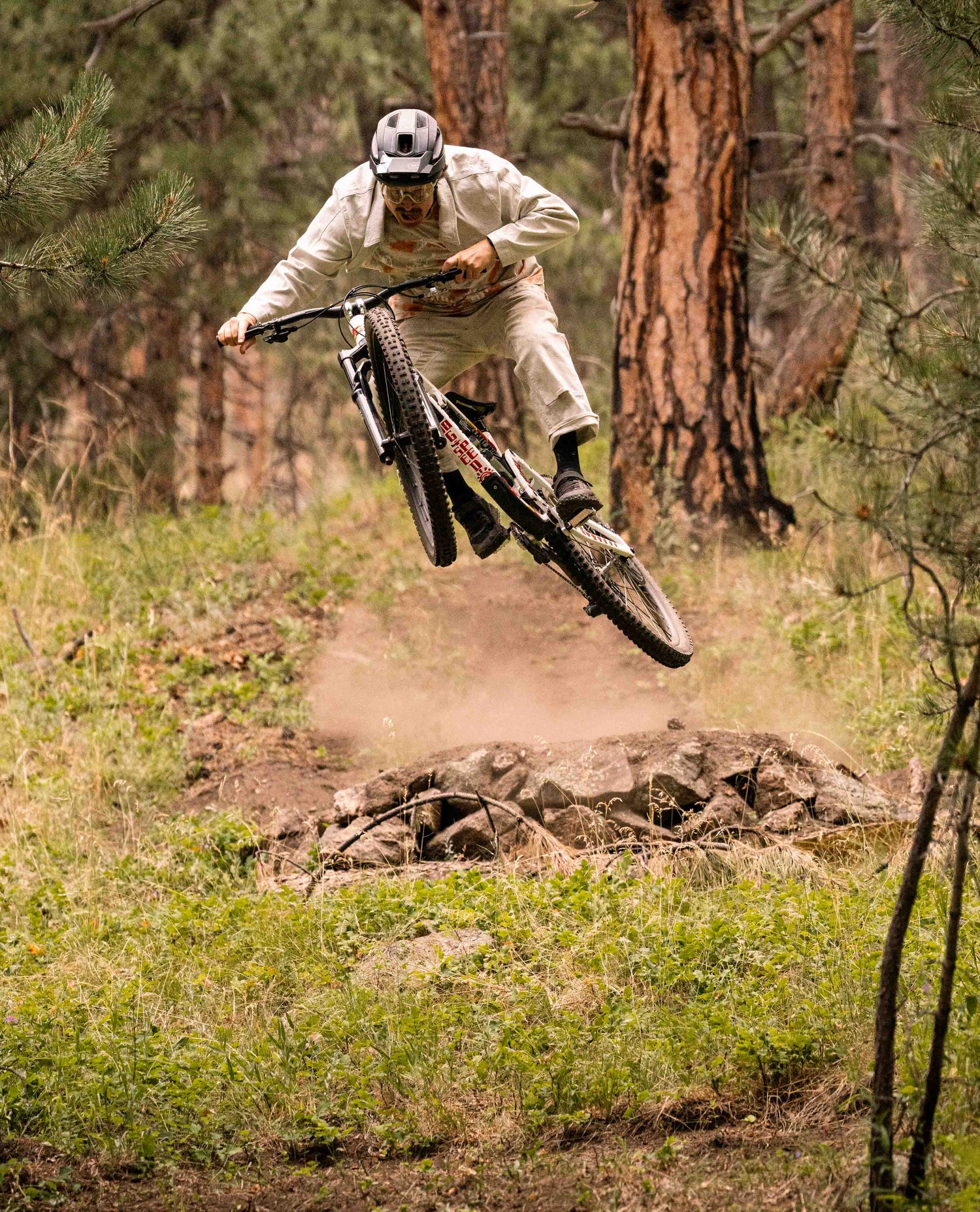 A man jumping his mountain bike in the woods. He is wearing a helmet, glasses, white jacket and pants, and a white and orange speckled t shirt. His bike is white with graffiti. 