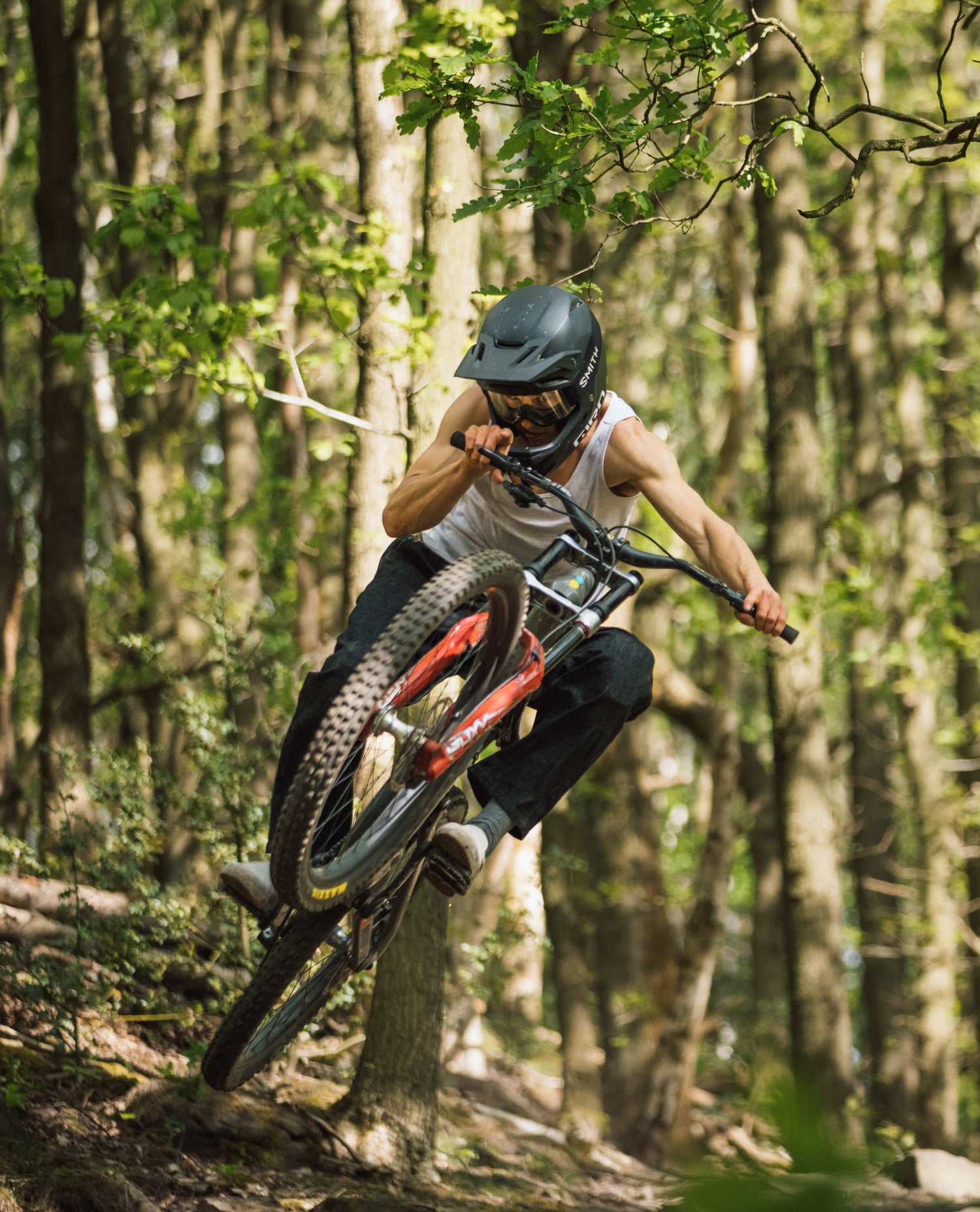 Man in white undershirt and black helmet jumping a mountain bike. 