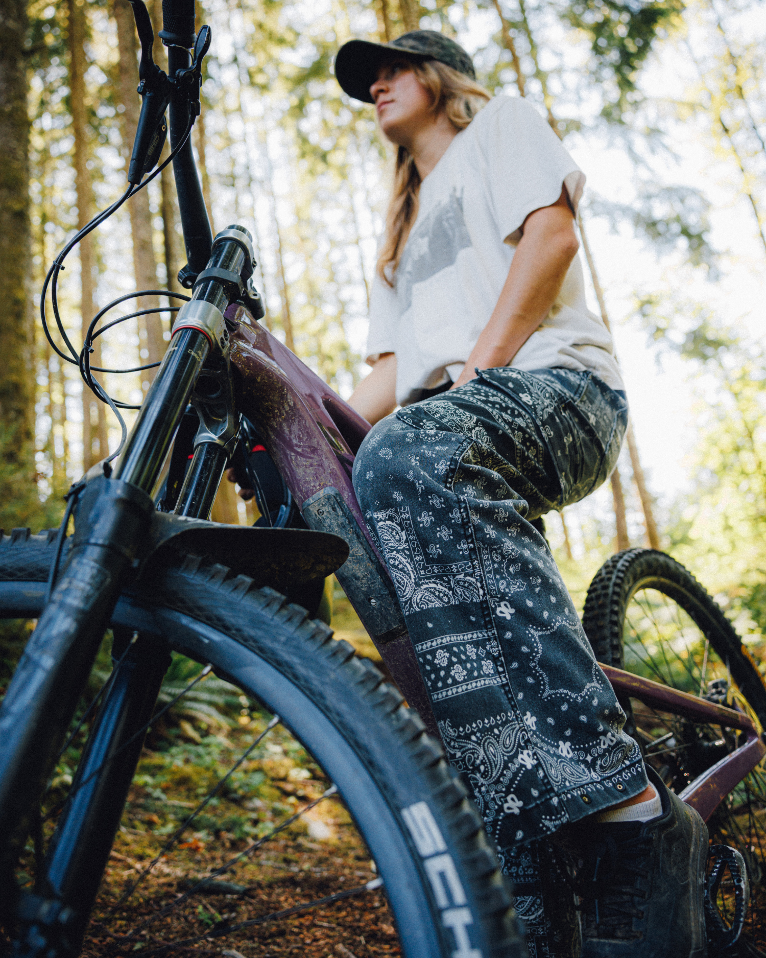 Woman wearing black bandana-print cargo pants, hat, and white shirt while sitting on a mountain bike in the forest.