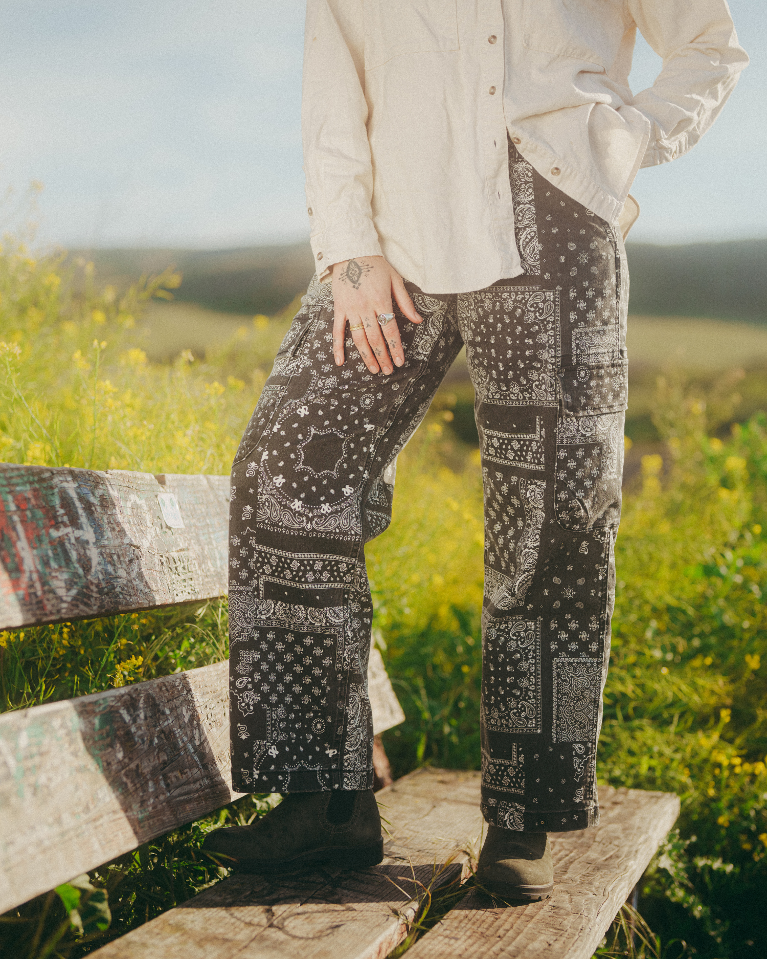 Black-and-white bandana-print cargo pants styled with a cream button-down shirt. Woman standing on wooden bench with green meadow in background