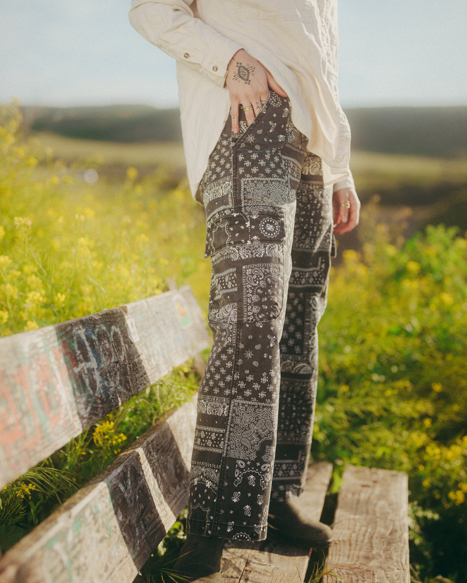 Close-up of black-and-white bandana-print cargo pants on a wooden bench outdoors with green meadow in background