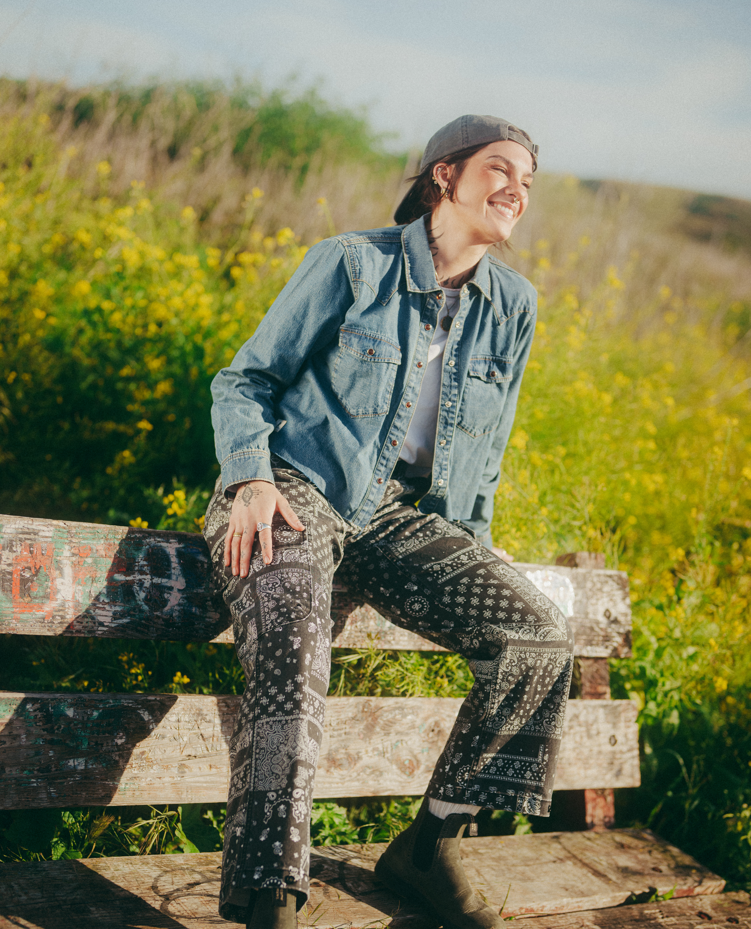 A person sitting on a weathered wooden bench outdoors in a field of yellow flowers, wearing a blue denim shirt, a gray hat, and  patterned bandana pants.