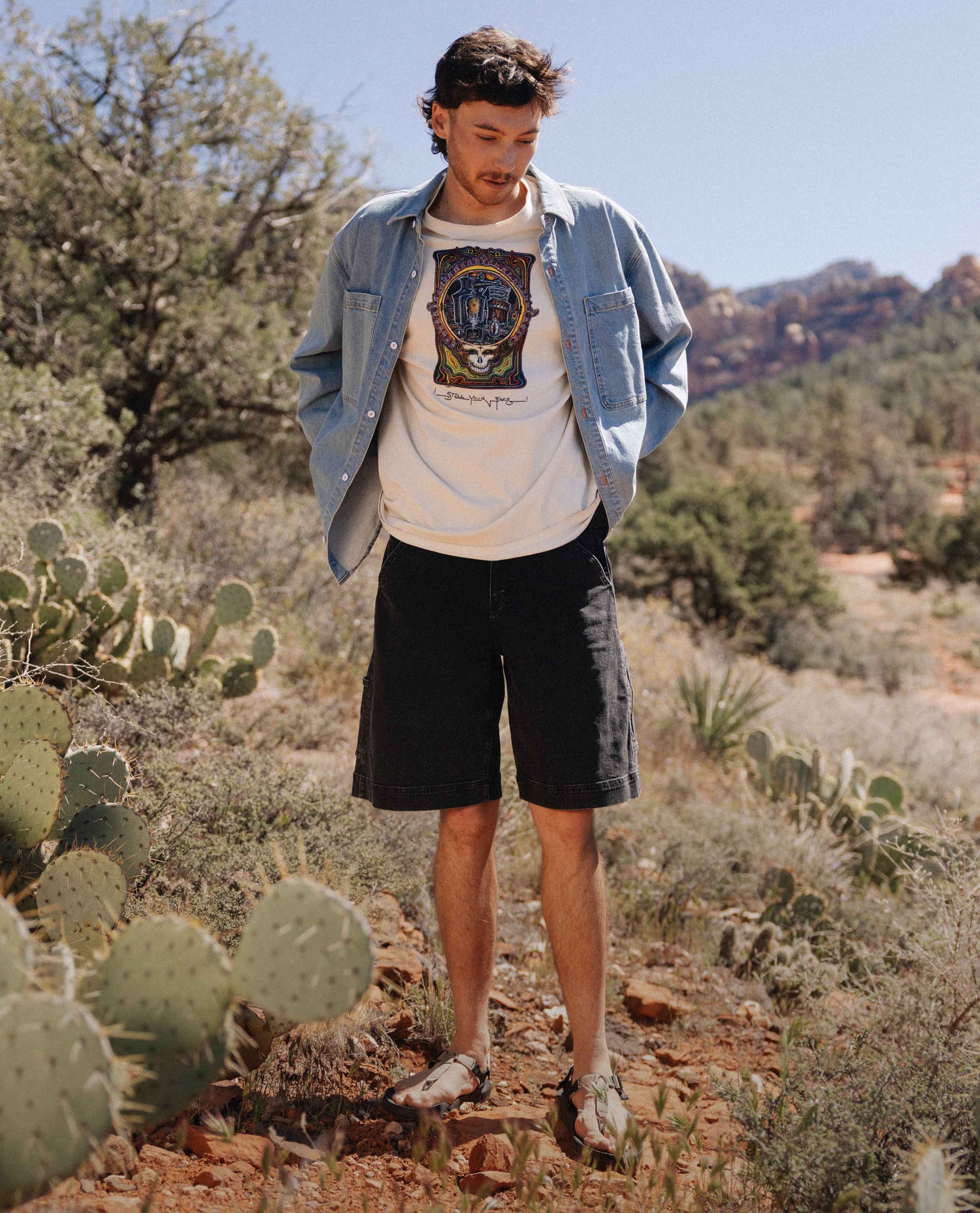 Man standing in a desert landscape wearing a denim jacket, graphic t-shirt, and black shorts.