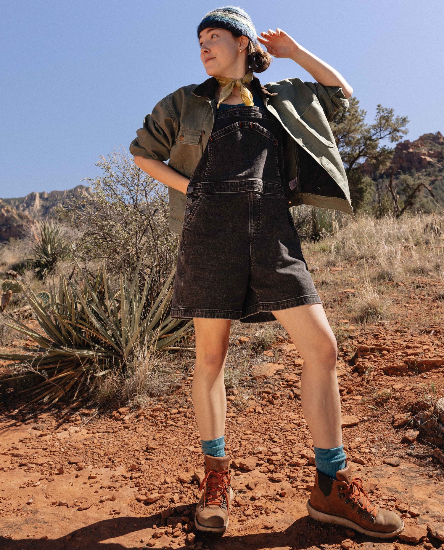 Person wearing a green jacket and black overalls standing in a desert landscape.