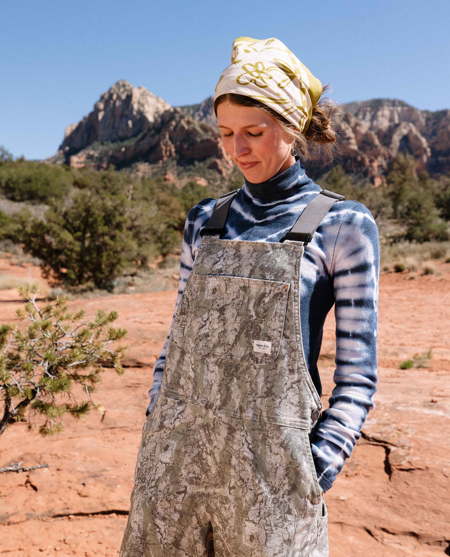 Person wearing a patterned apron and headscarf in a desert landscape with mountains.