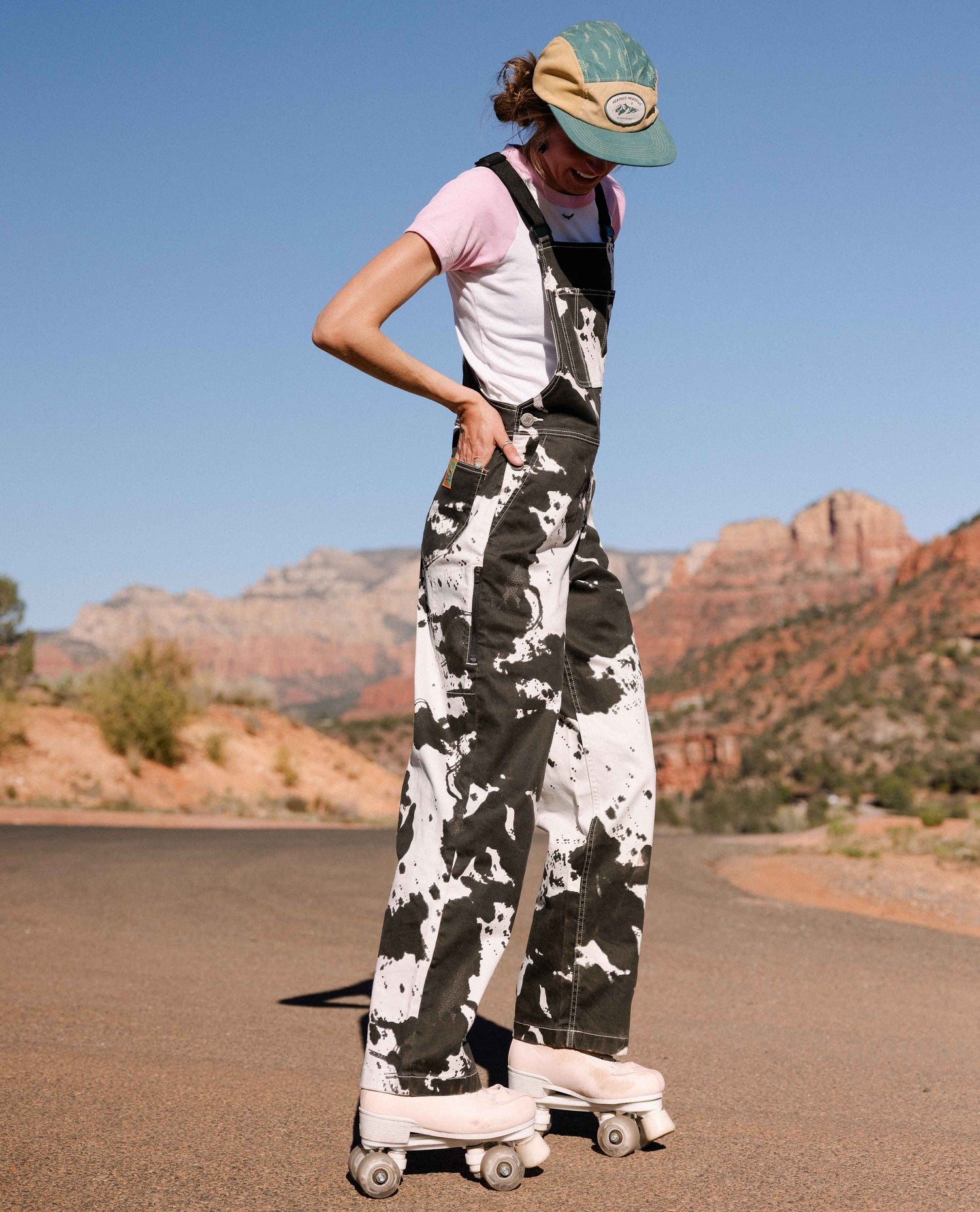 Person wearing a pink shirt, white and black patterned overalls, and a cap standing on roller skates with a desert landscape in the background.