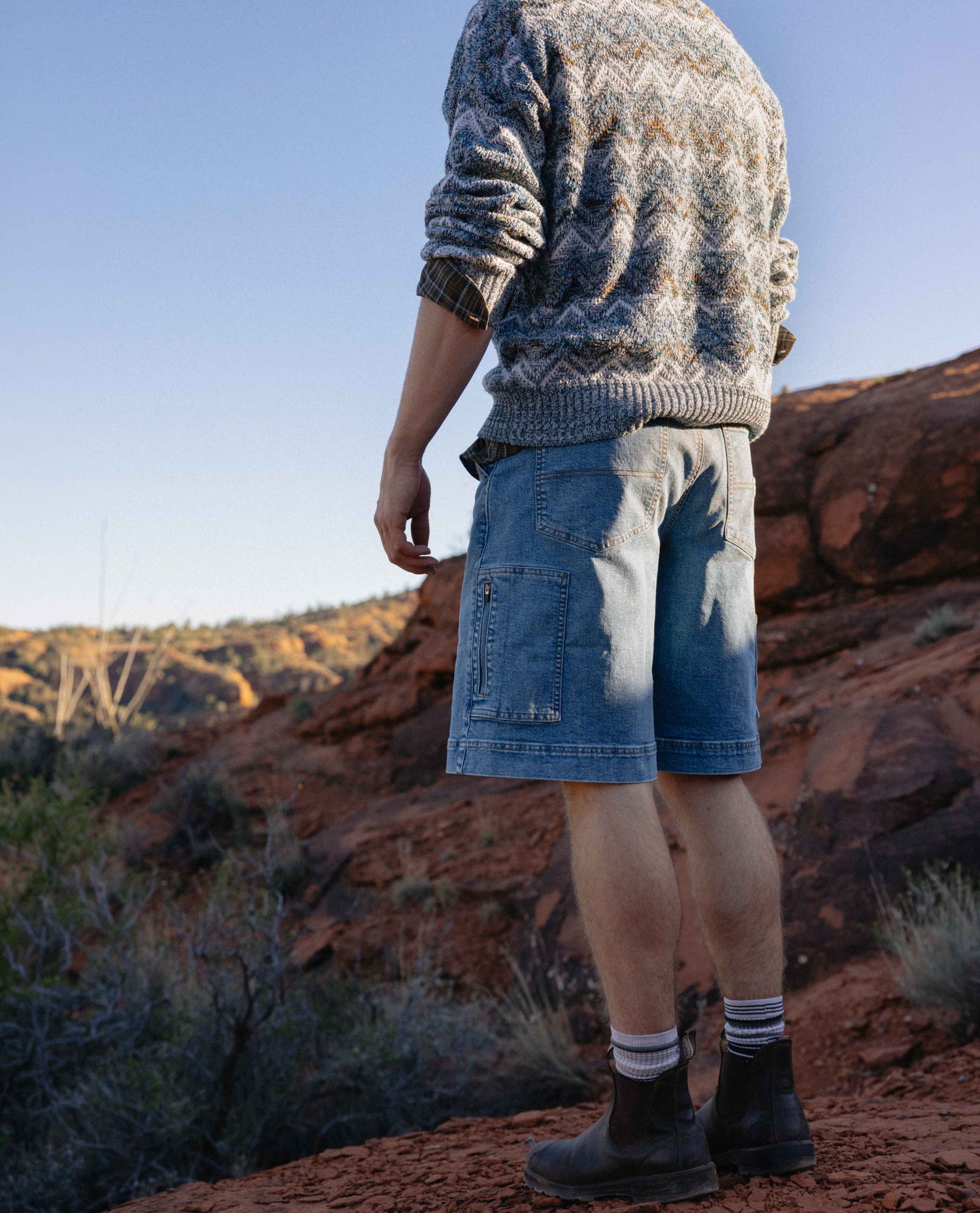 Person wearing a patterned sweater and denim shorts standing in a desert landscape.