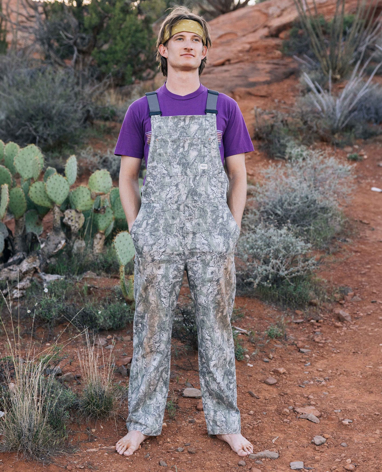 Person wearing camouflage overalls and a purple shirt in a desert setting with cacti and rocks.