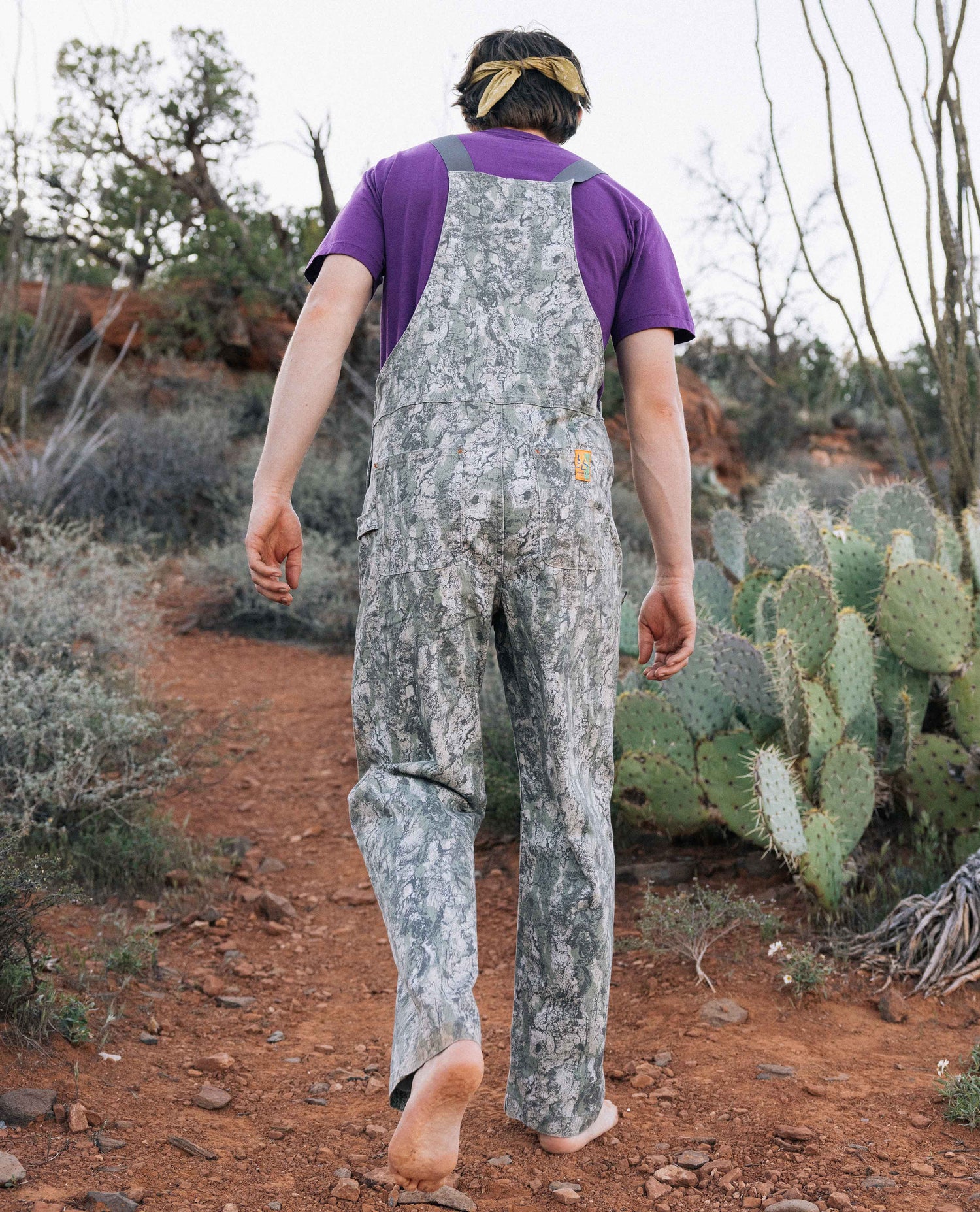 Person wearing camouflage overalls and a purple shirt walking on a dirt path with cacti in the background