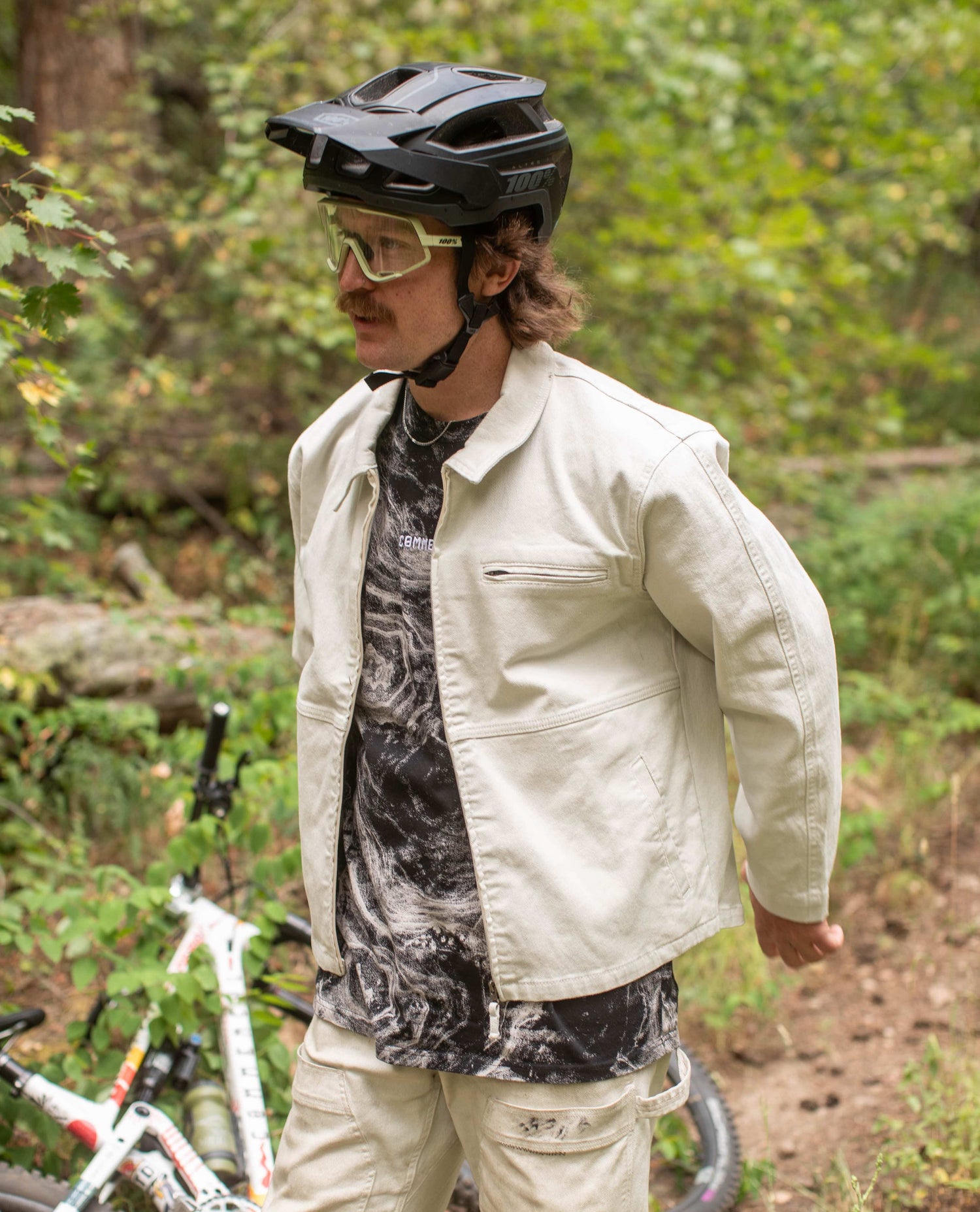 A man wearing a mountain bike helmet, glasses, a white jacket with matching white pants, and a mottled grey and black jersey. A mountain bike lays behind him out of focus. 