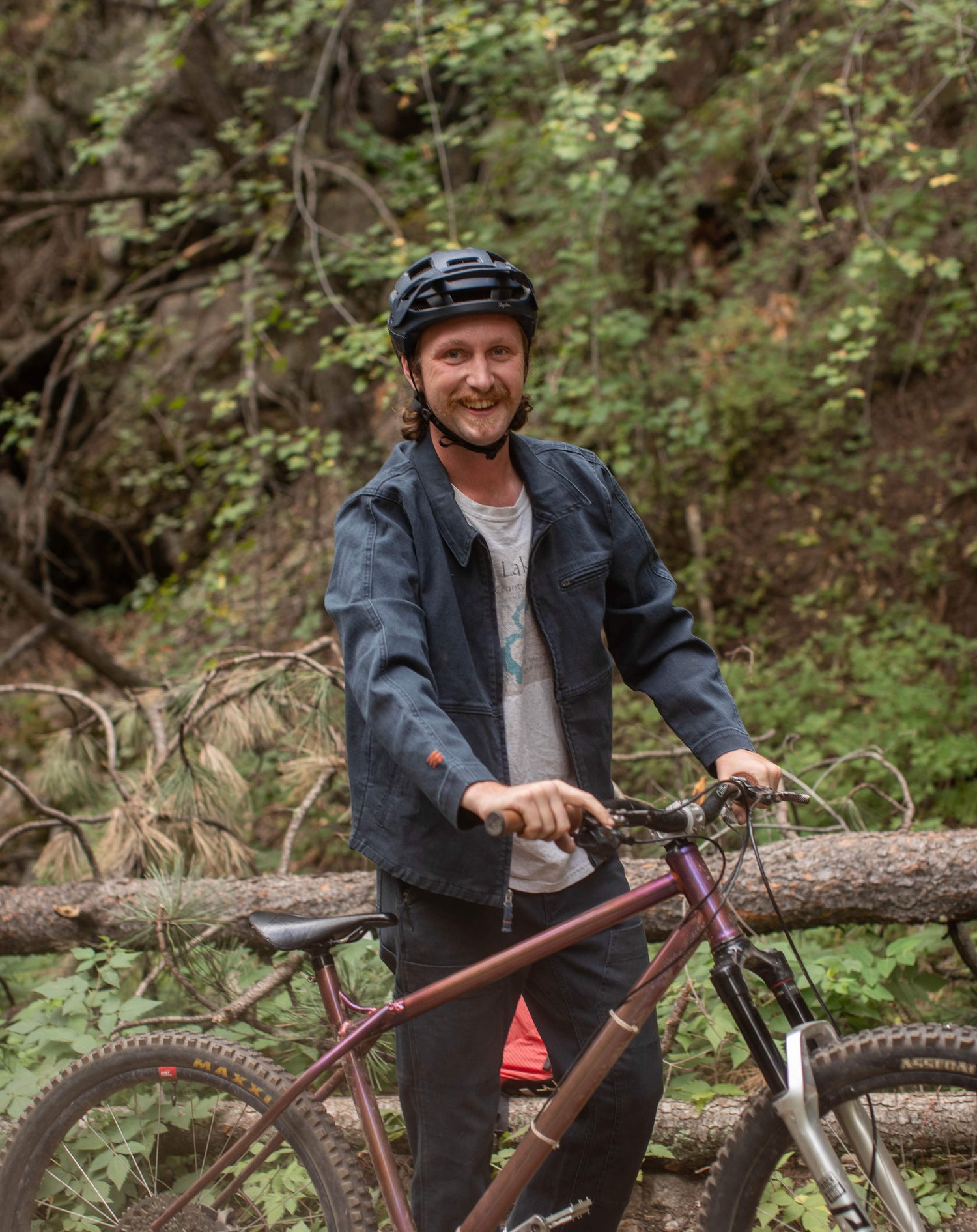 A man with a mustache and a mullet smiling at camera. He is pushing a mountain bike and is wearing a blue helmet, blue jacket, and blue cargo pants. 