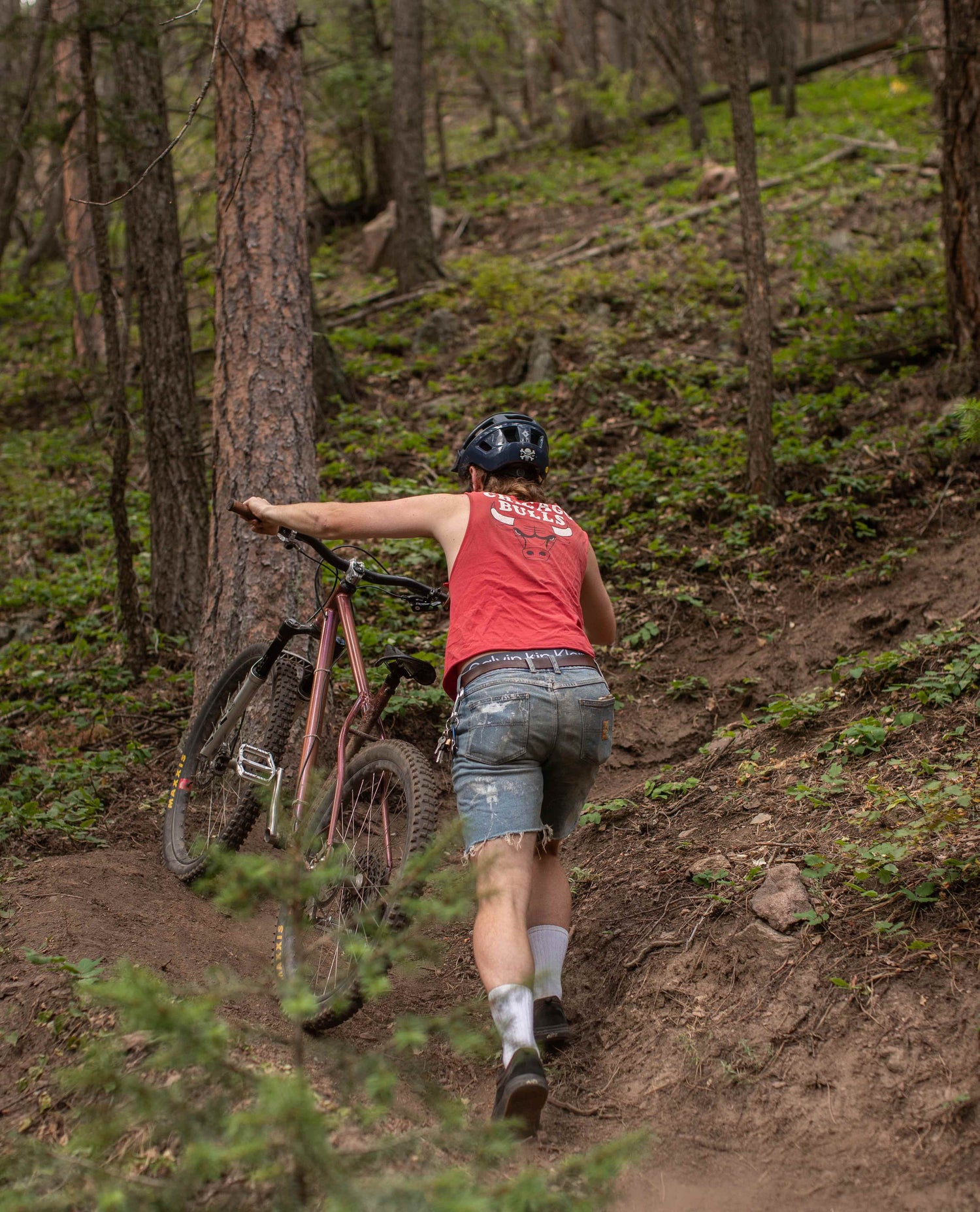 A man sitting on his mountain bike on a trail in the woods. He is wearing a red "BULLS" tank top and worn and paint splattered blue jean shorts. He has tall white socks. 