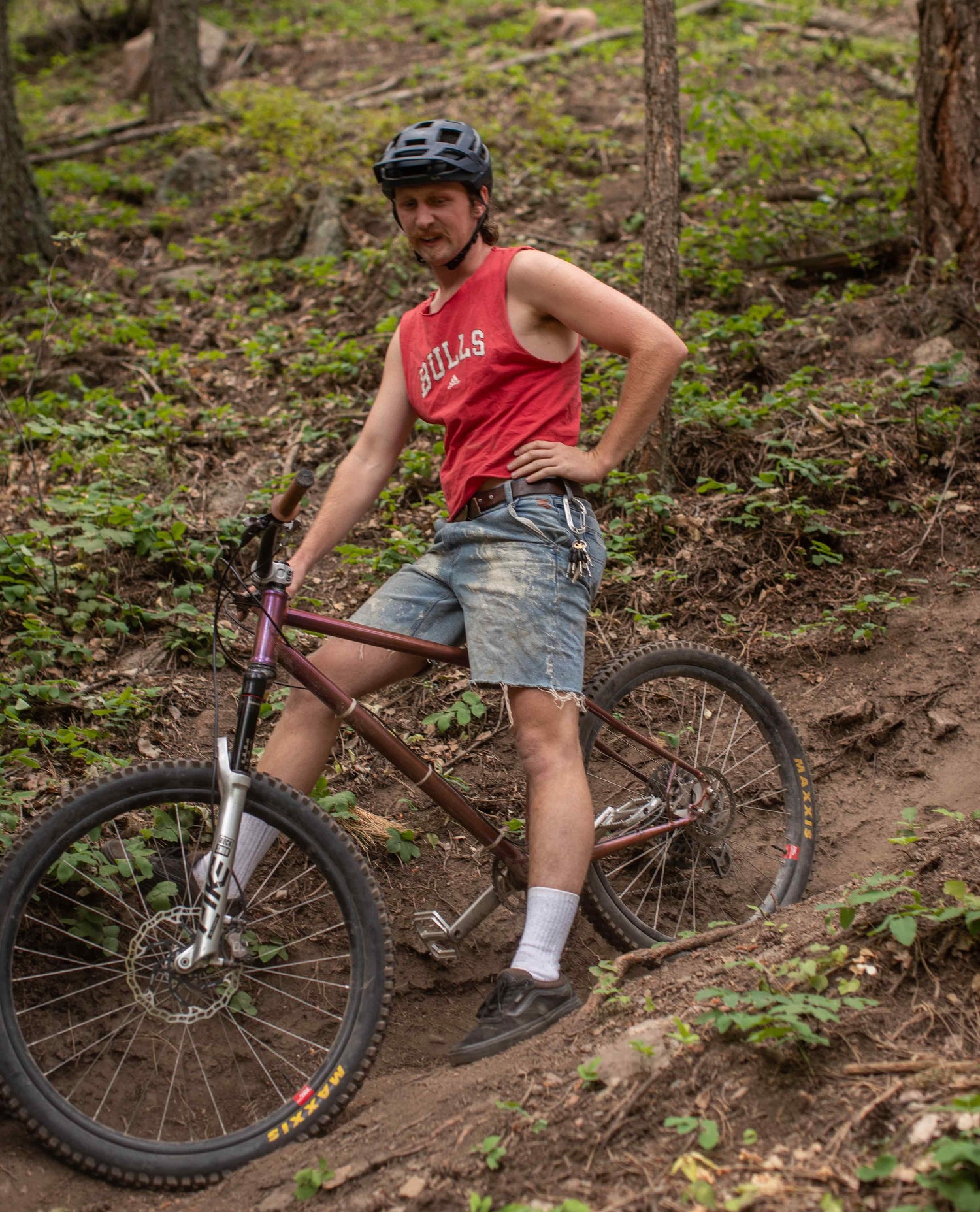 A man sitting on his mountain bike on a trail in the woods. He is wearing a red "BULLS" tank top and worn and paint splattered blue jean shorts. He has tall white socks. 