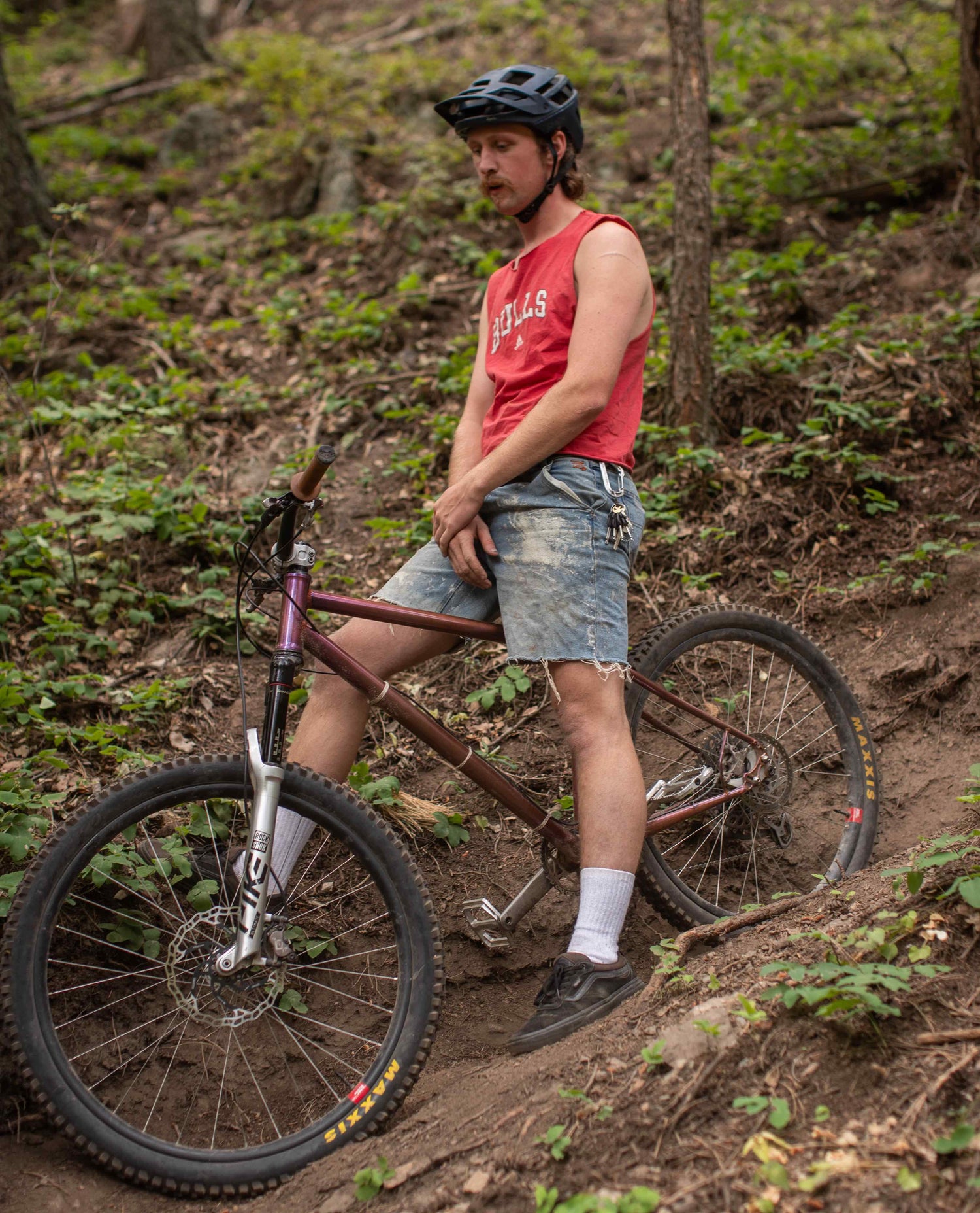 A man sitting on his mountain bike on a trail in the woods. He is wearing a red "BULLS" tank top and worn and paint splattered blue jean shorts. He has tall white socks. 