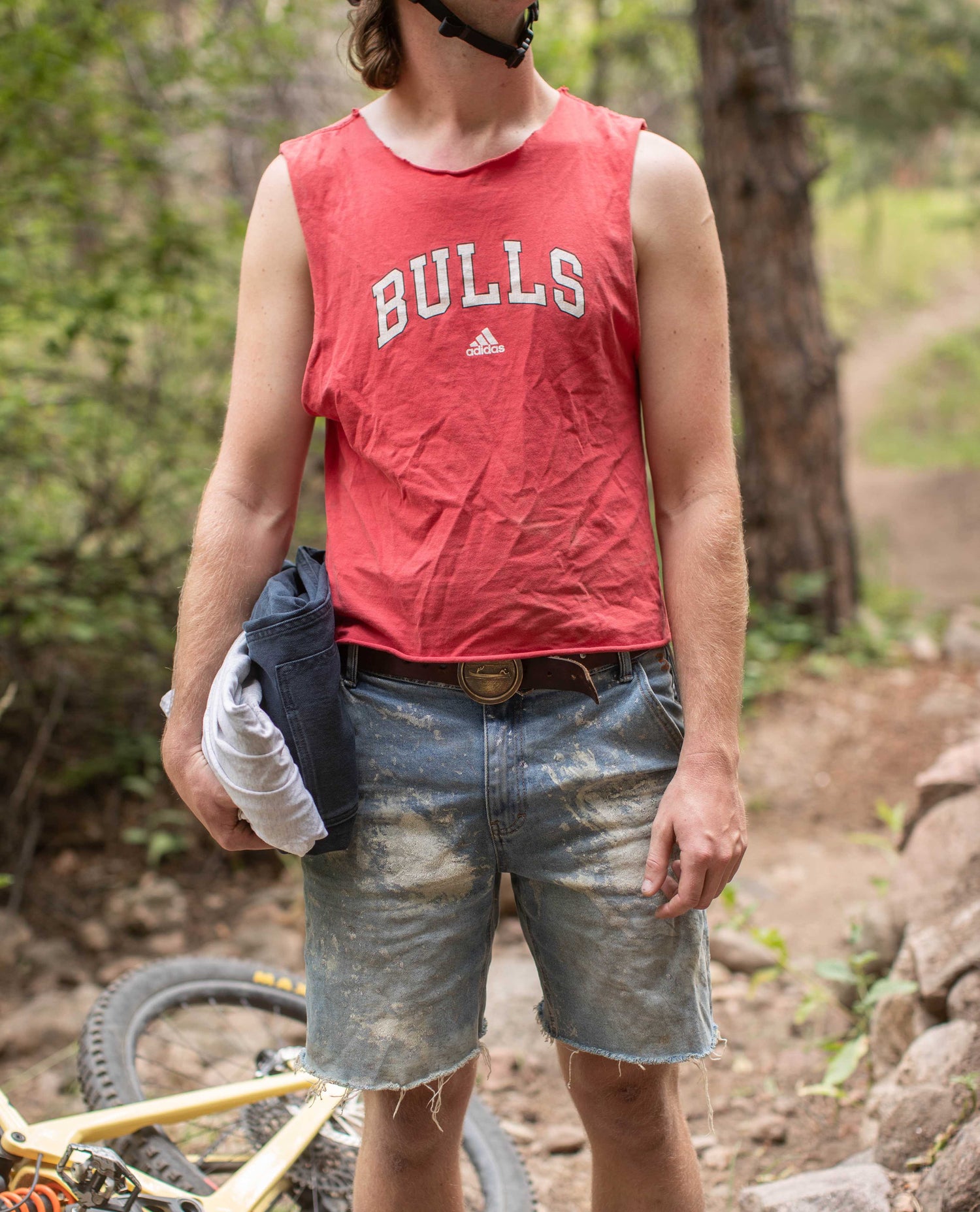 A man in a red "BULLS" tank top and blue worn out jean shorts stands in front of camera. His face is not visible. He is carrying additional clothing in his right hand. A mountain bike leans out of focus to his right. 