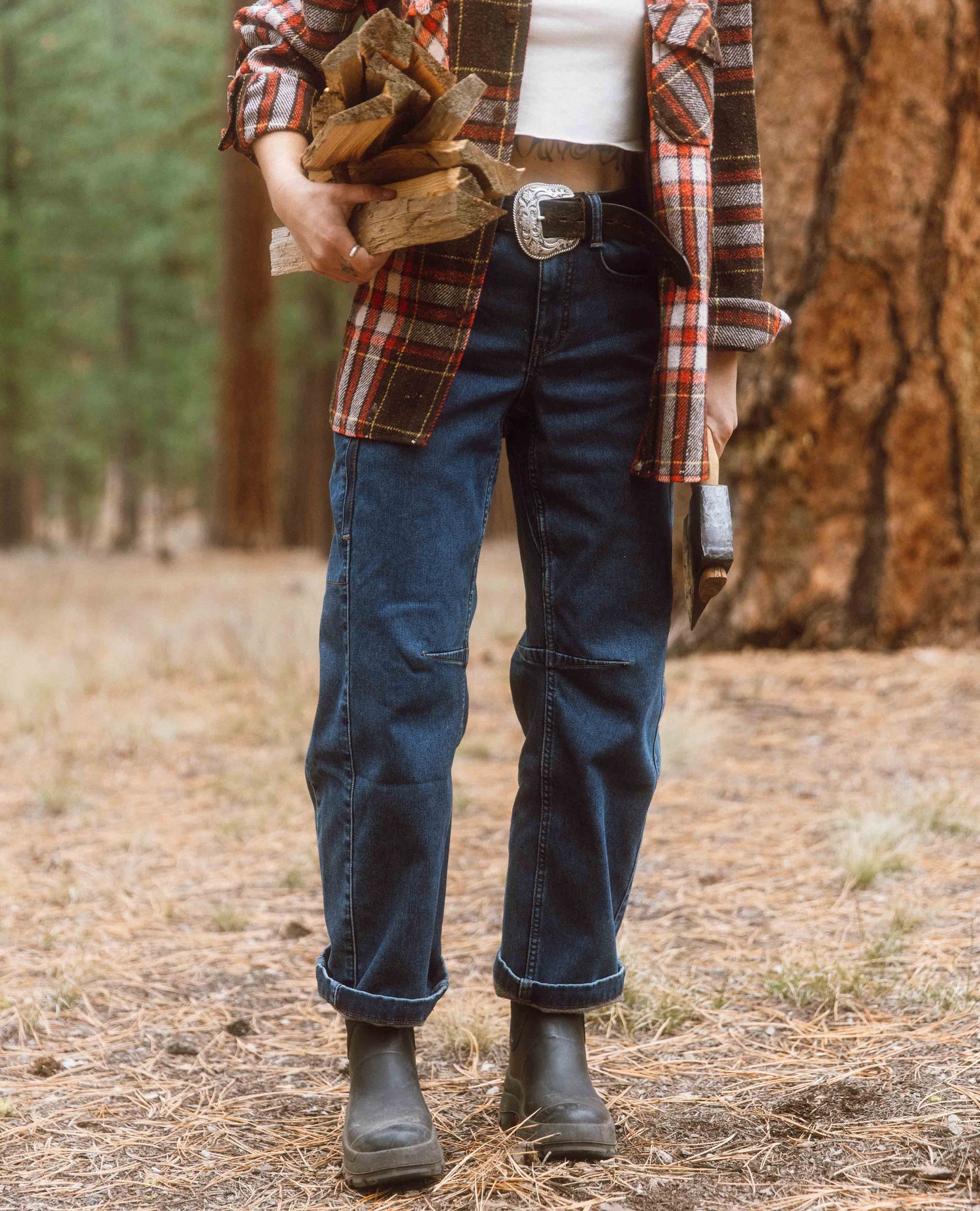 Person holding firewood in a forest wearing a plaid shirt, jeans, and boots.