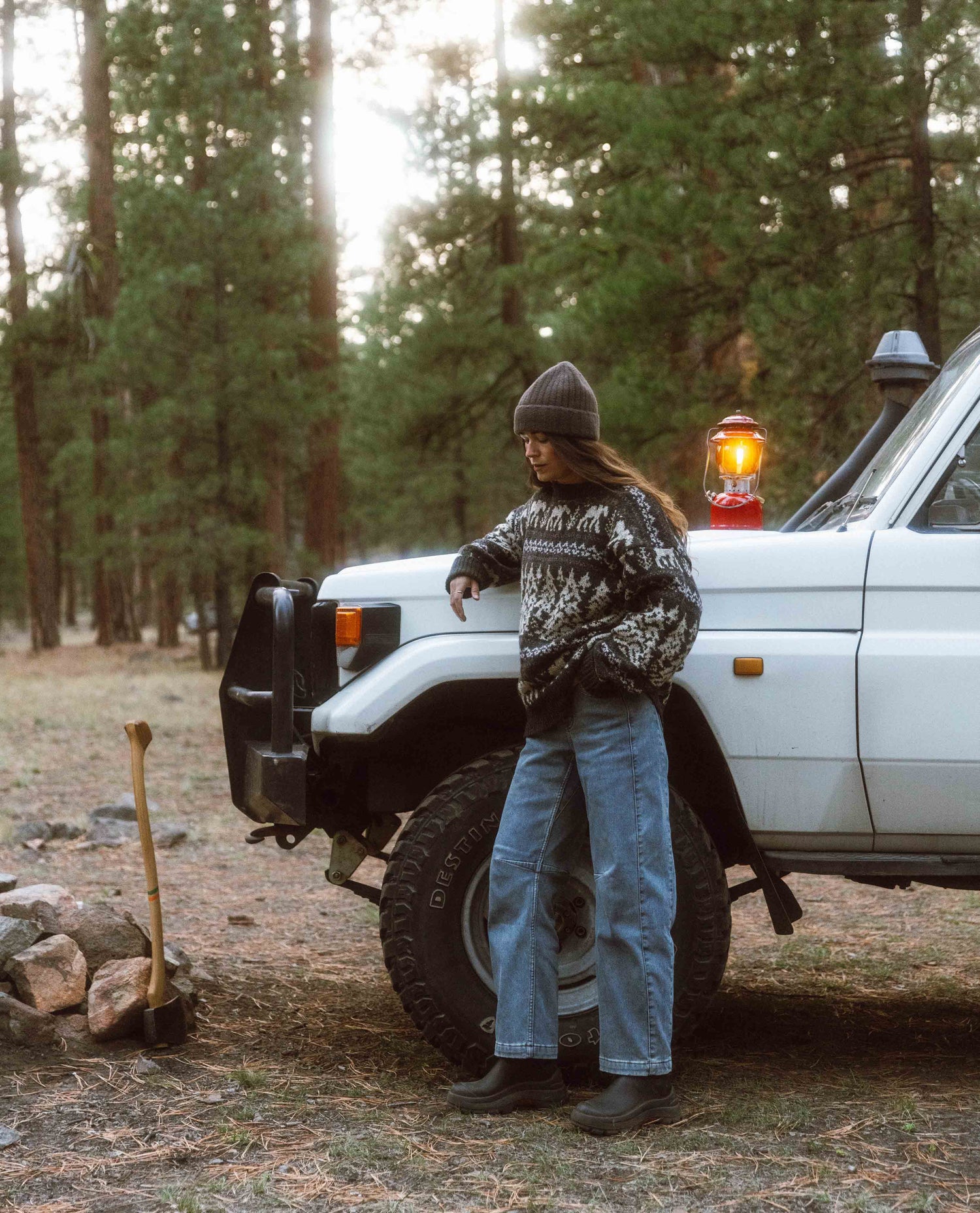 Person standing next to a white truck in a forest setting with a lantern on the hood.