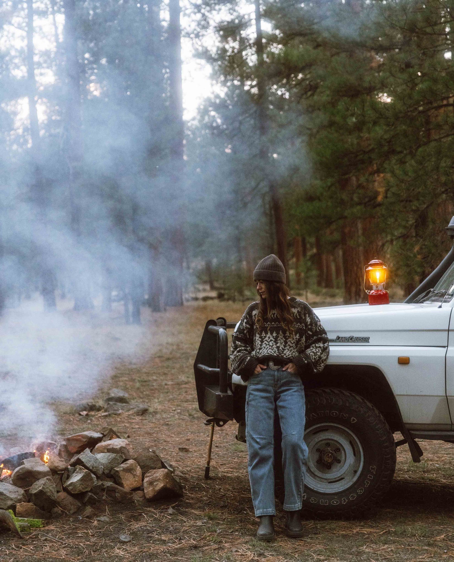 Person standing by a campfire and vehicle in a forest setting