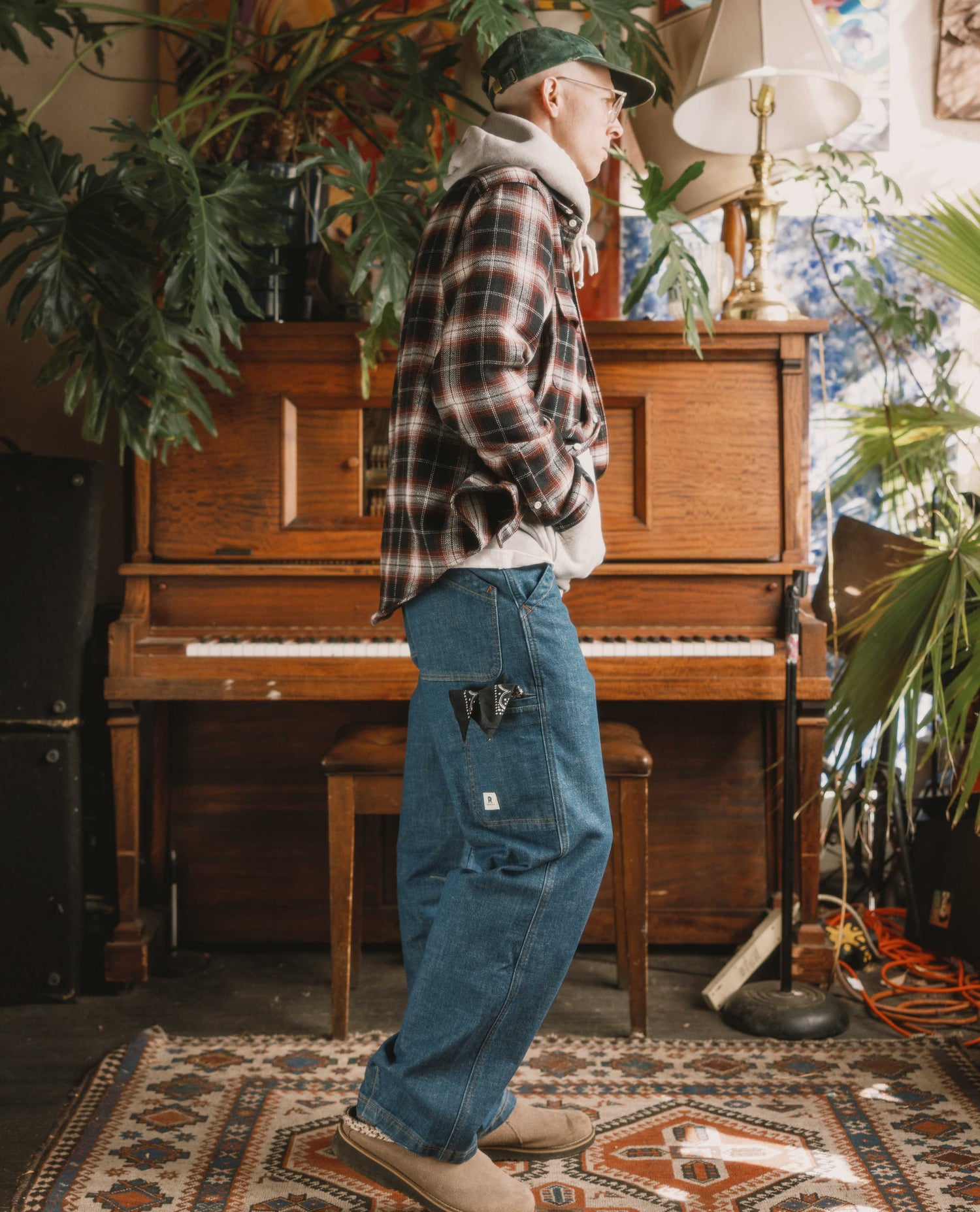 Person wearing a plaid shirt and jeans standing in front of a wooden piano with plants around.