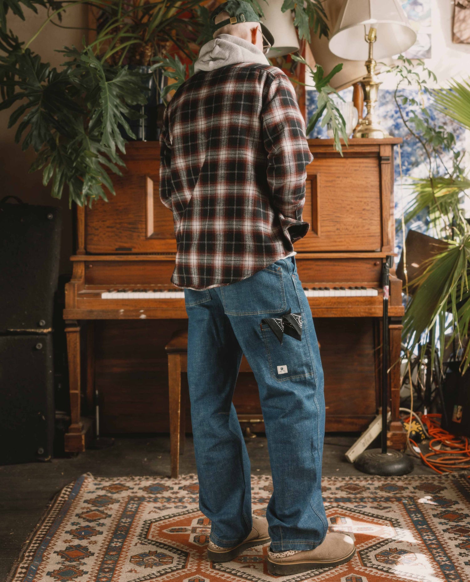 Person wearing a plaid shirt and blue jeans standing in a room with a piano and plants.